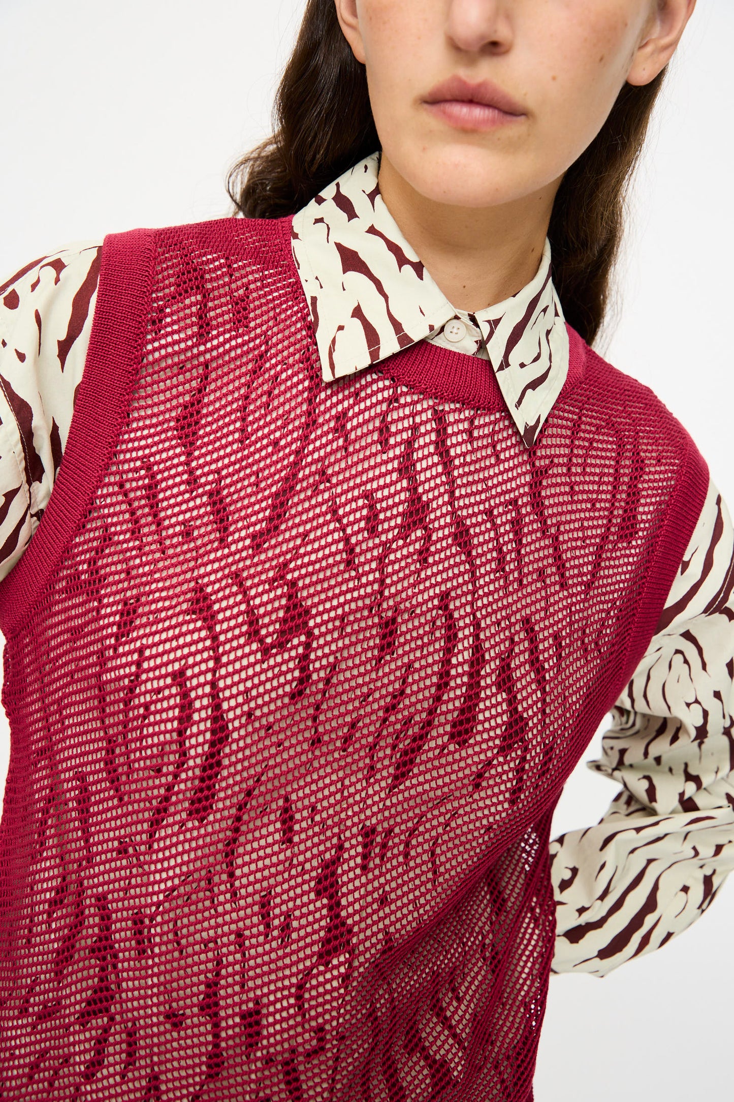A woman wears the Rachel Comey Rhett Tank in Deep Red, a sleeveless, textured mesh sweater vest styled over a cream and burgundy patterned shirt, blending modern design with classic elements.