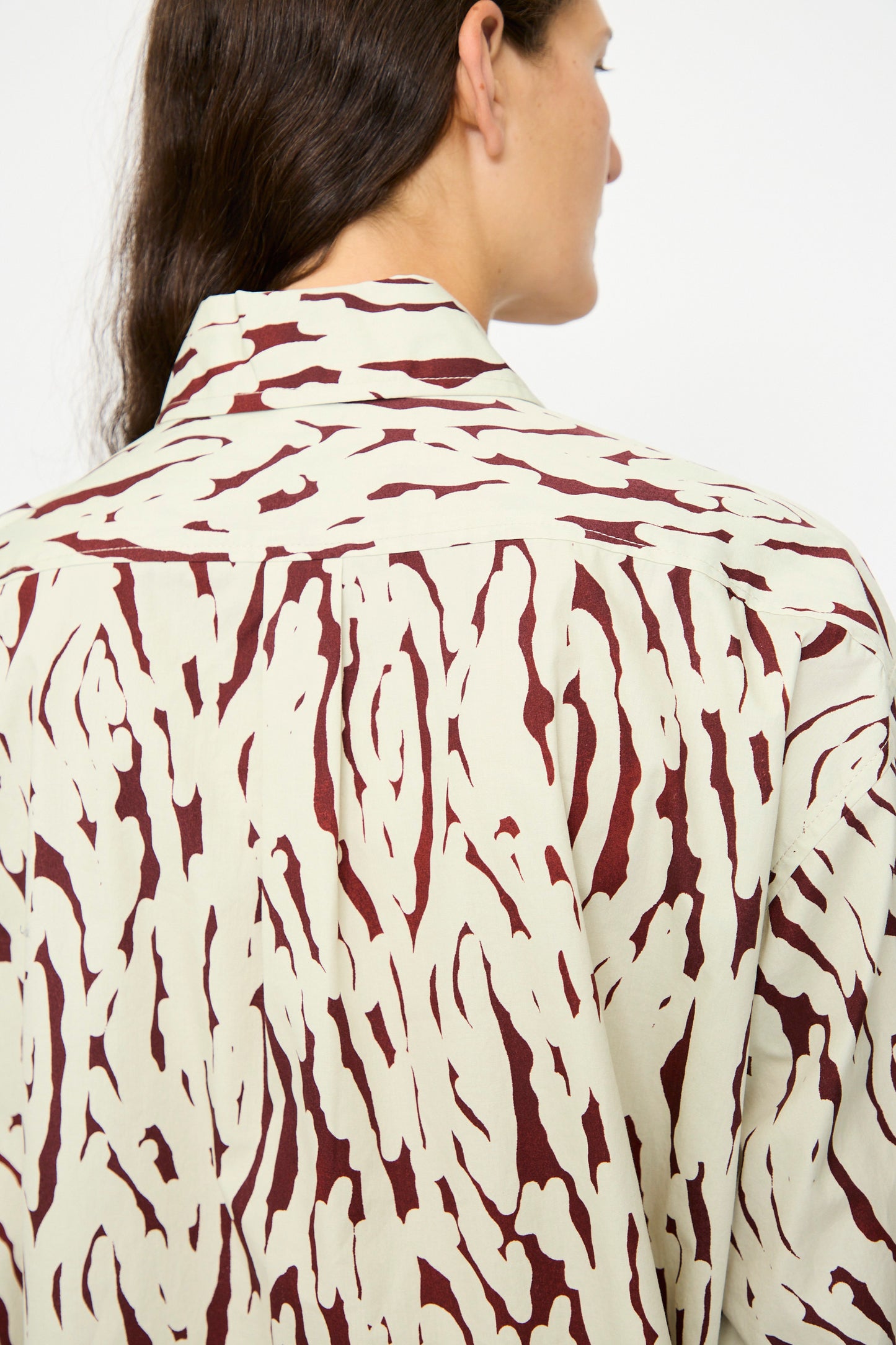 A person with long brown hair models the Risa Top in Cream by Rachel Comey, an oversized button-down featuring an abstract brown pattern, shown from the back and side.