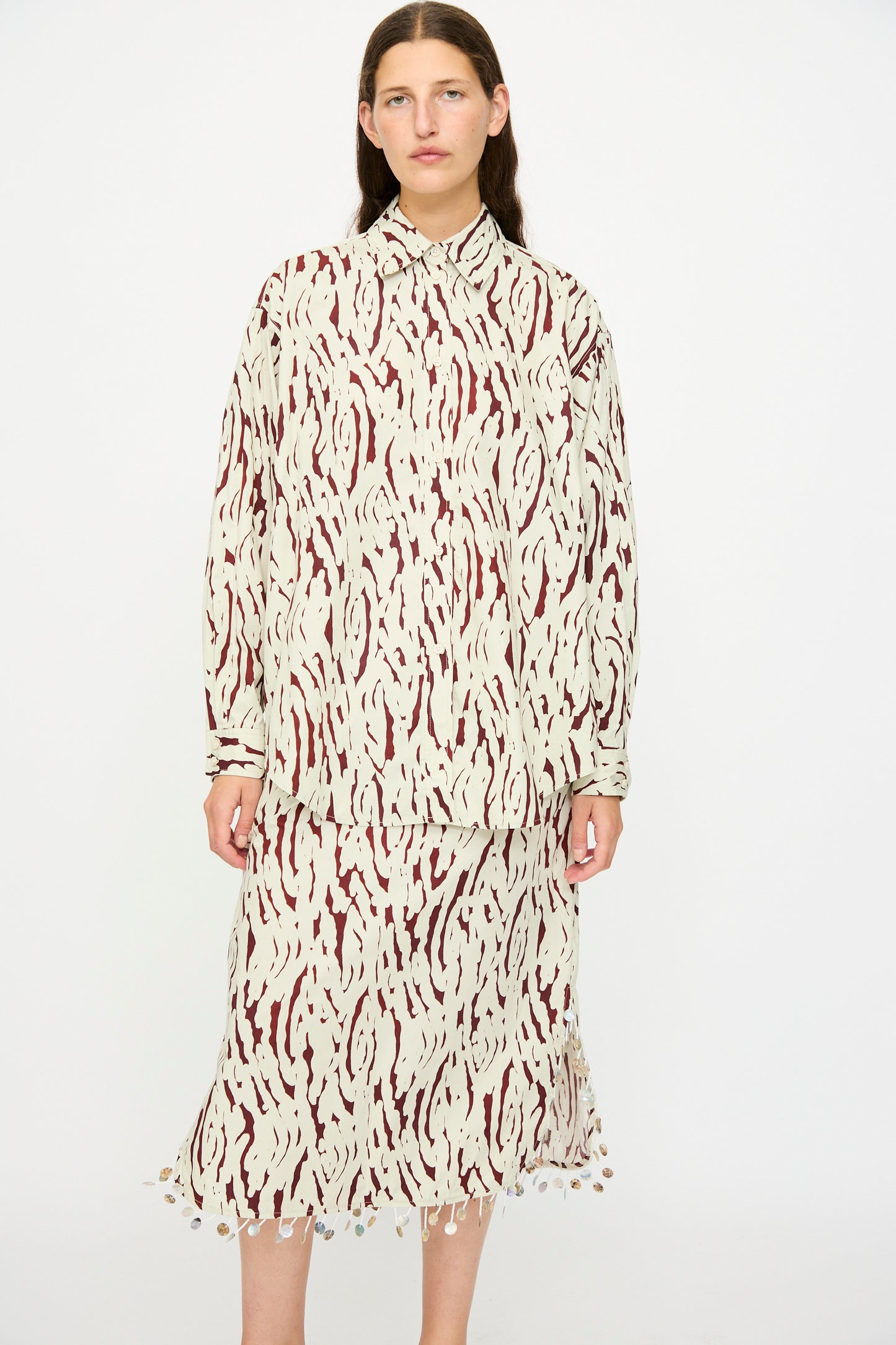 A woman stands against a plain background wearing the Rachel Comey Risa Top in Cream, an oversized button-down shirt featuring an abstract beige and maroon pattern.