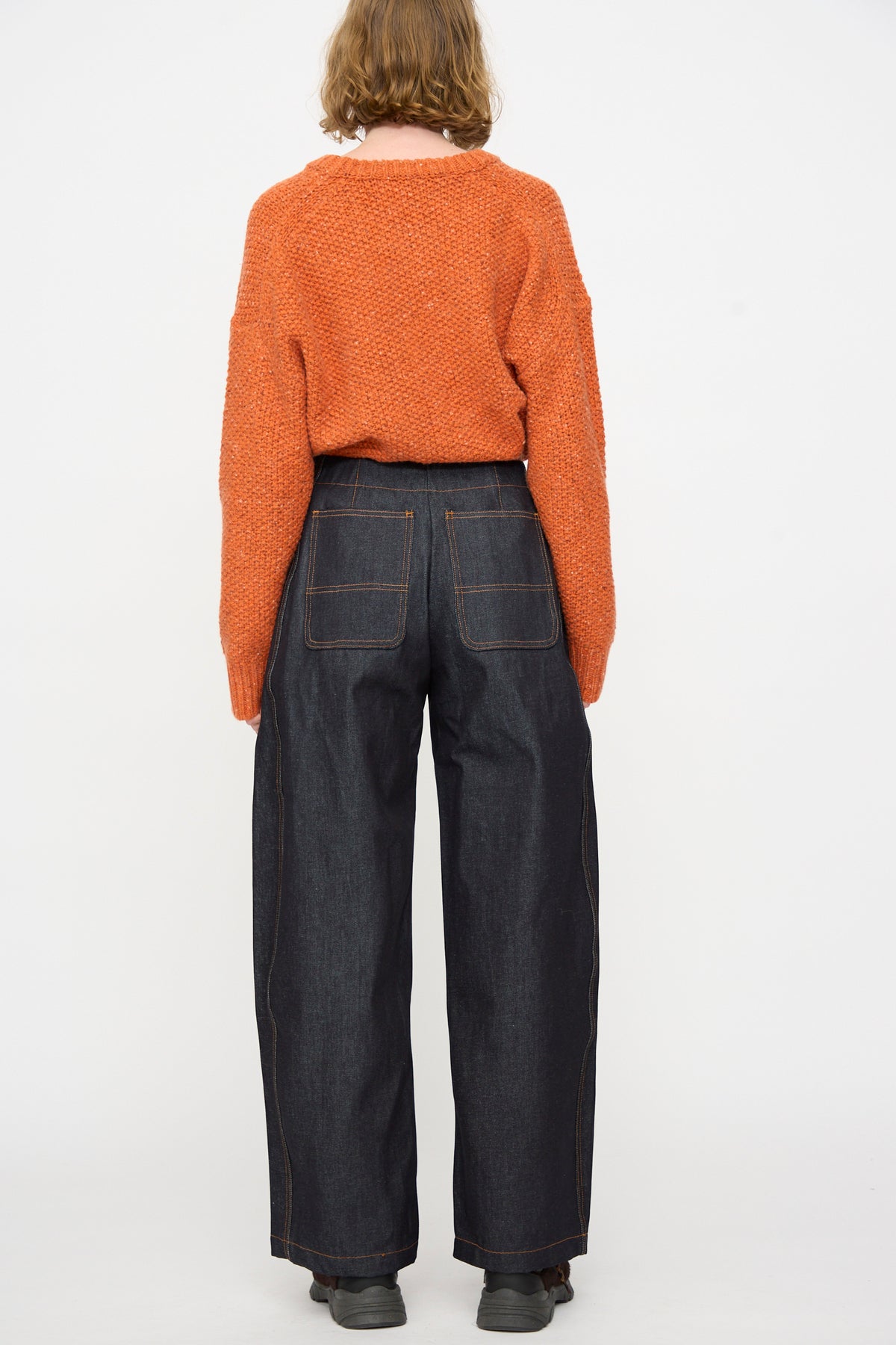 Person standing with back to camera, wearing an orange sweater and the Rachel Comey Tandy Pant in Super Raw, styled with gray shoes against a plain white background.