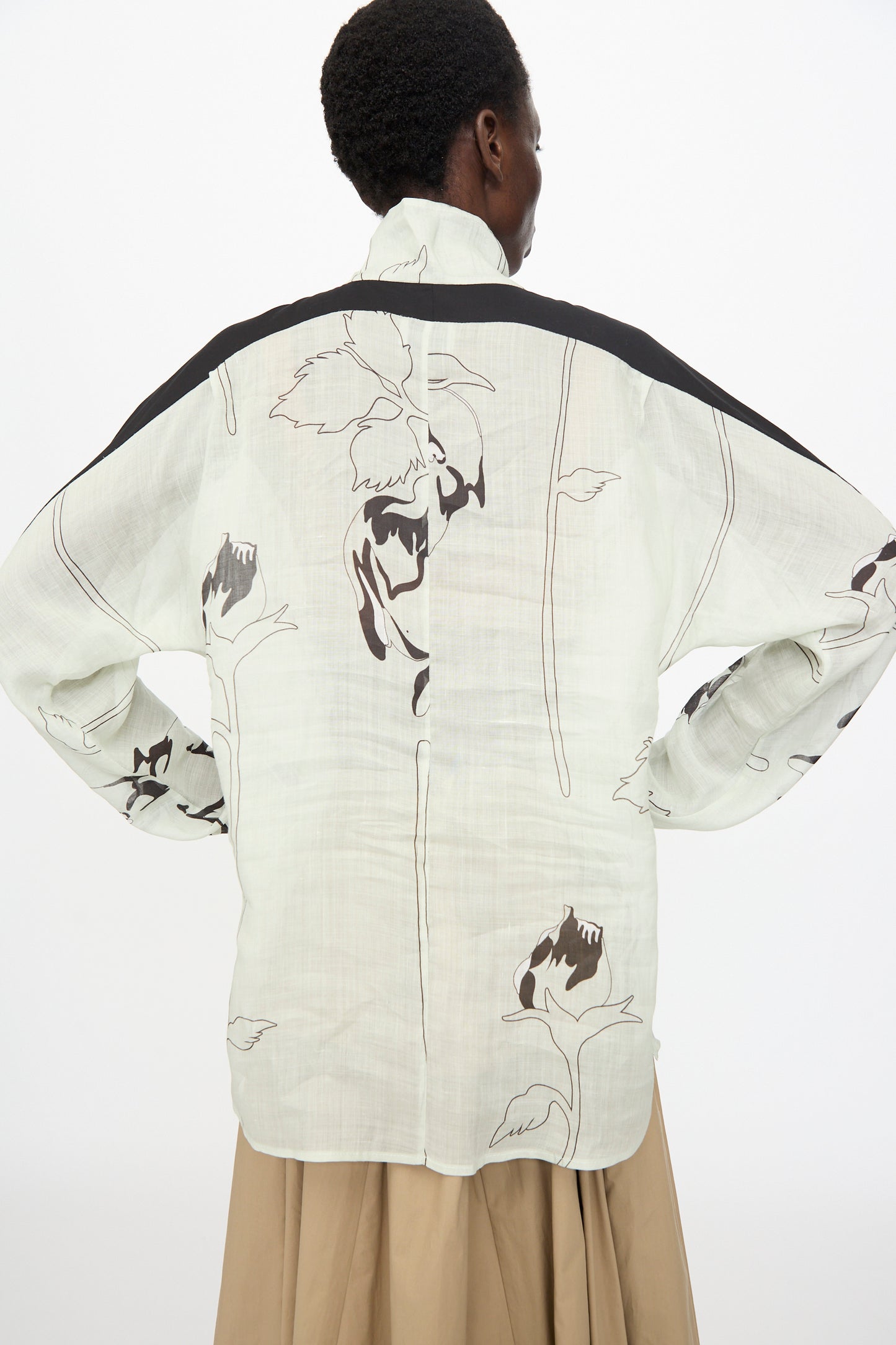 A person stands with their back to the camera, wearing the Rachel Comey Wanson Top in Dove—a light-colored, long sleeve shirt with a black floral print and shoulder stripe—paired with a beige skirt.