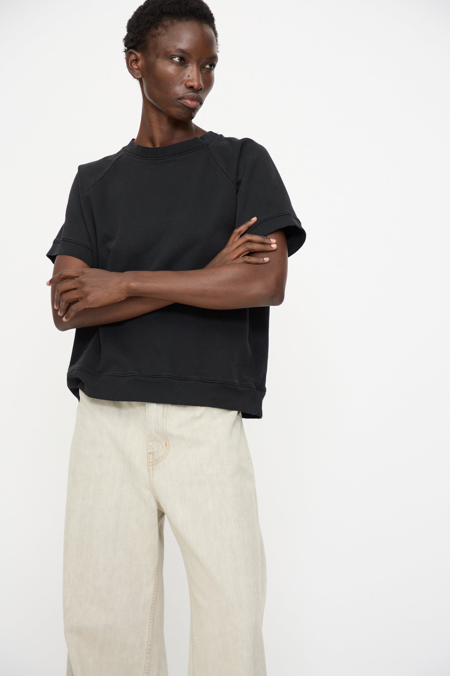 Person standing against a plain white background, arms crossed and neutral expression, wearing the Rachel Comey Billi Sweatshirt in Charcoal and light beige wide-leg pants.