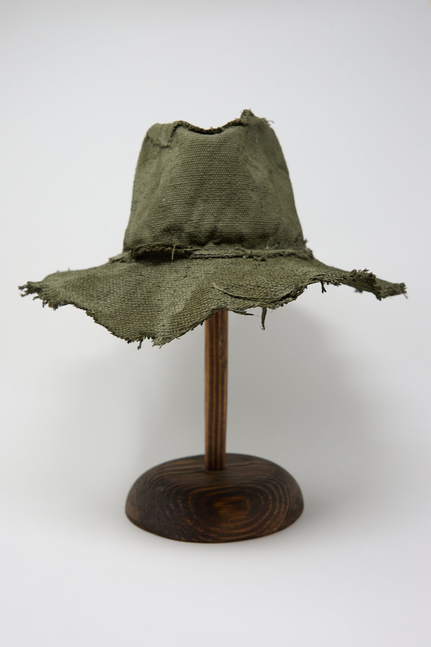 A worn and frayed Reinhard Plank Beghe S Jute Hat in Green displayed on a wooden stand against a white background.