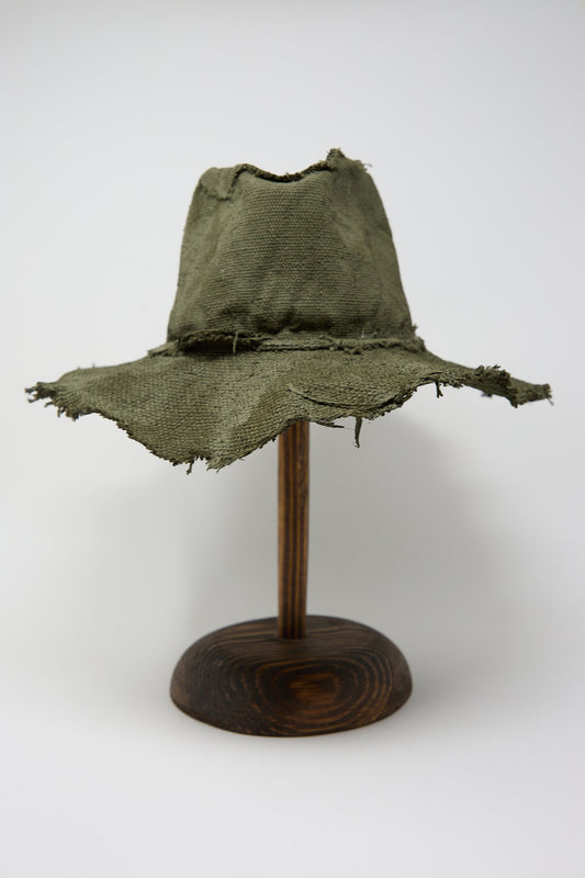 A worn and frayed Reinhard Plank Beghe S Jute Hat in Green displayed on a wooden stand against a white background.