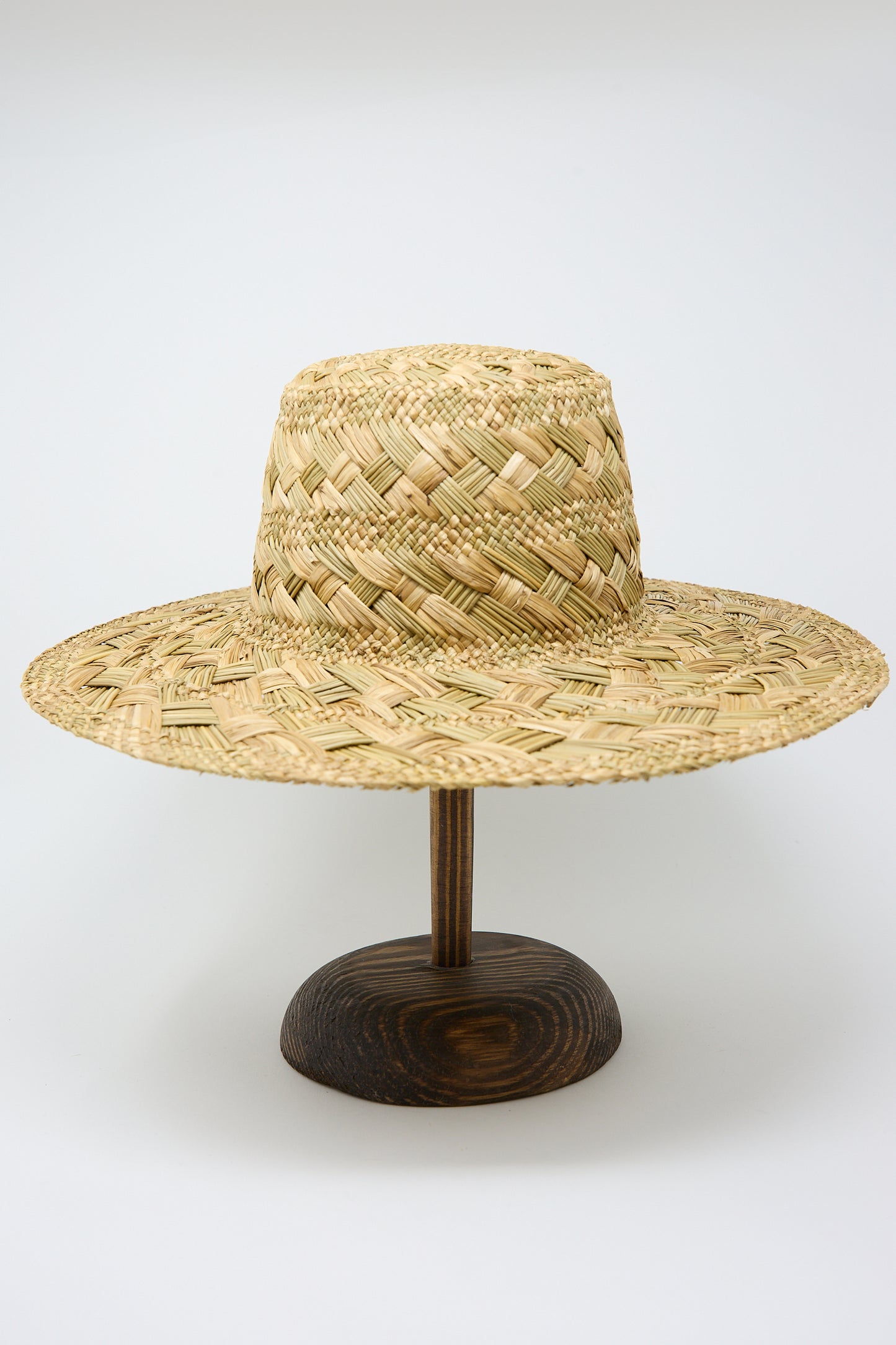 A wide-brimmed Treccia Lai Straw Hat in Natural displayed on a wooden stand against a plain white background by Reinhard Plank.