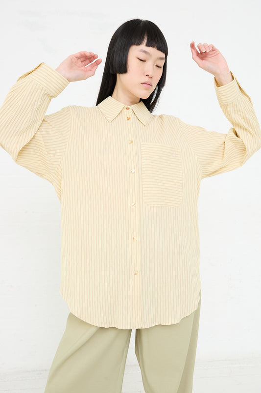 A woman with black hair, wearing a Rejina Pyo Organic Cotton Seersucker Caprice Shirt in Stripe Yellow and green trousers, stands with her eyes closed and arms raised slightly.
