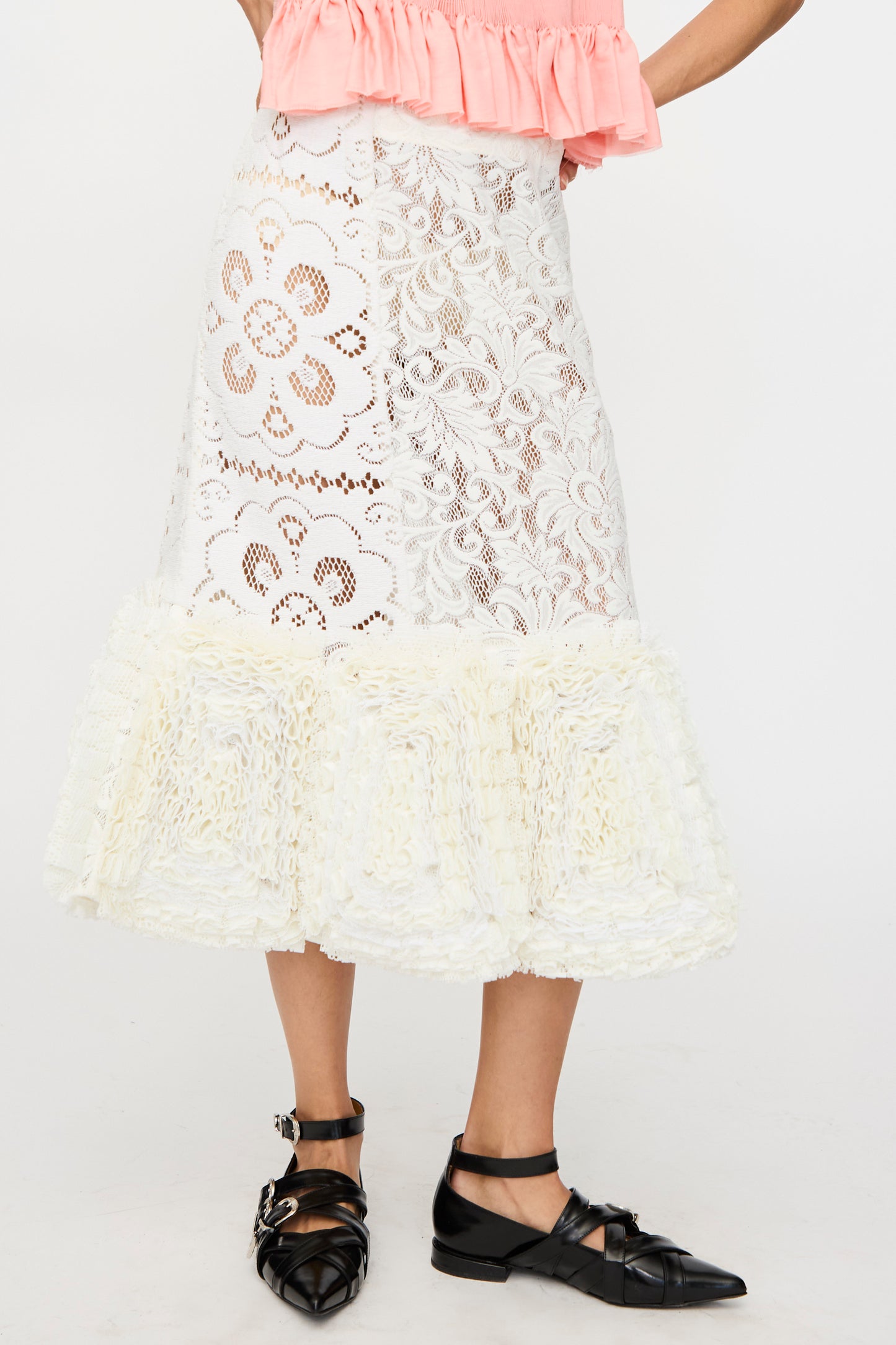 A person wears the Acarajé Skirt in Tonal White by Renata Brenha, featuring white lace with floral patterns and a ruffled hem, paired with a pink off-shoulder top and black pointed-toe buckled shoes.