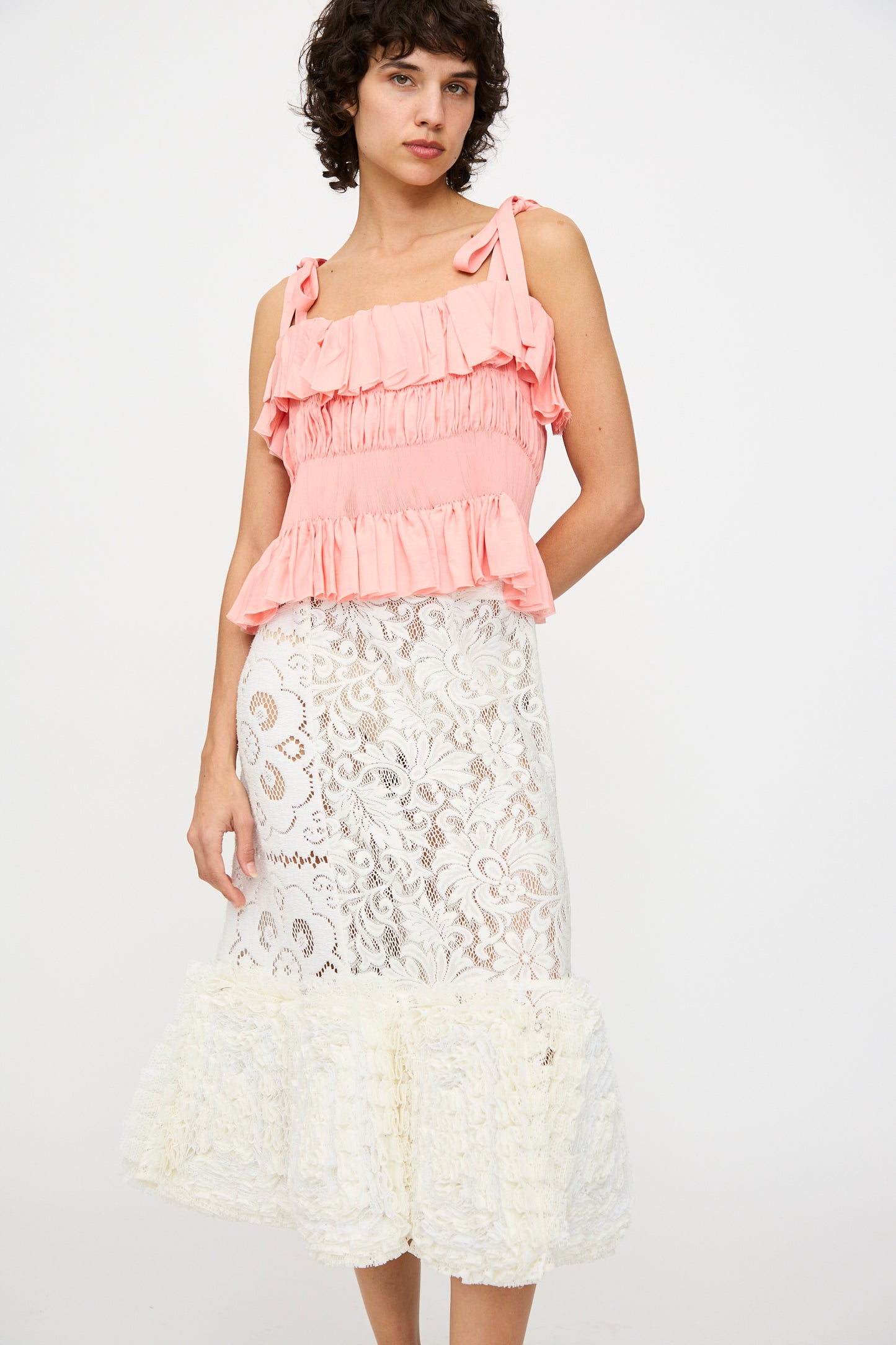 A person wearing a sleeveless pink ruffled top and the Acarajé Skirt in Tonal White by Renata Brenha, featuring a ruffled hem made with the frufru technique, stands against a plain white background.