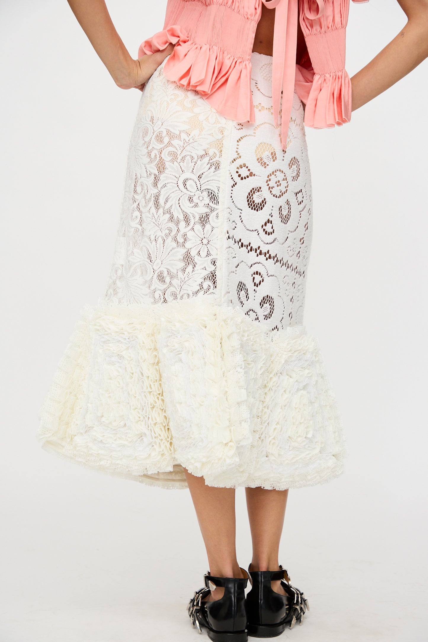A person wears the Acarajé Skirt in Tonal White by Renata Brenha, a white lace and textured midi skirt made from up-cycled curtains using the frufru technique, styled with a pink ruffled top and black studded flats.