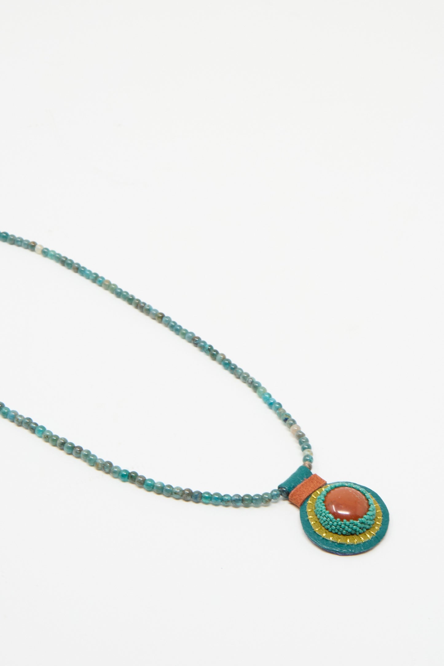 The Charm Necklace by Robin Mollicone features handmade apatite beads with a circular pendant adorned with a red jasper stone, set against a white background.