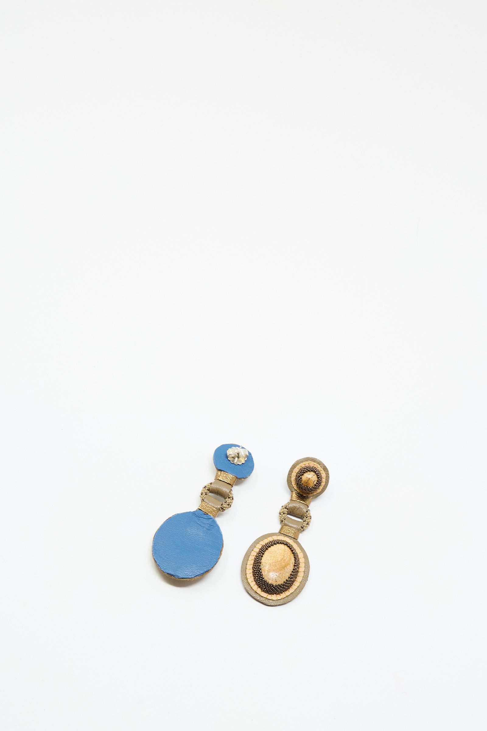 Two mismatched Robin Mollicone earrings on a white background: one is the Double Stone Earrings in Picture Jasper, round with blue double stones; the other is gold featuring concentric oval shapes.