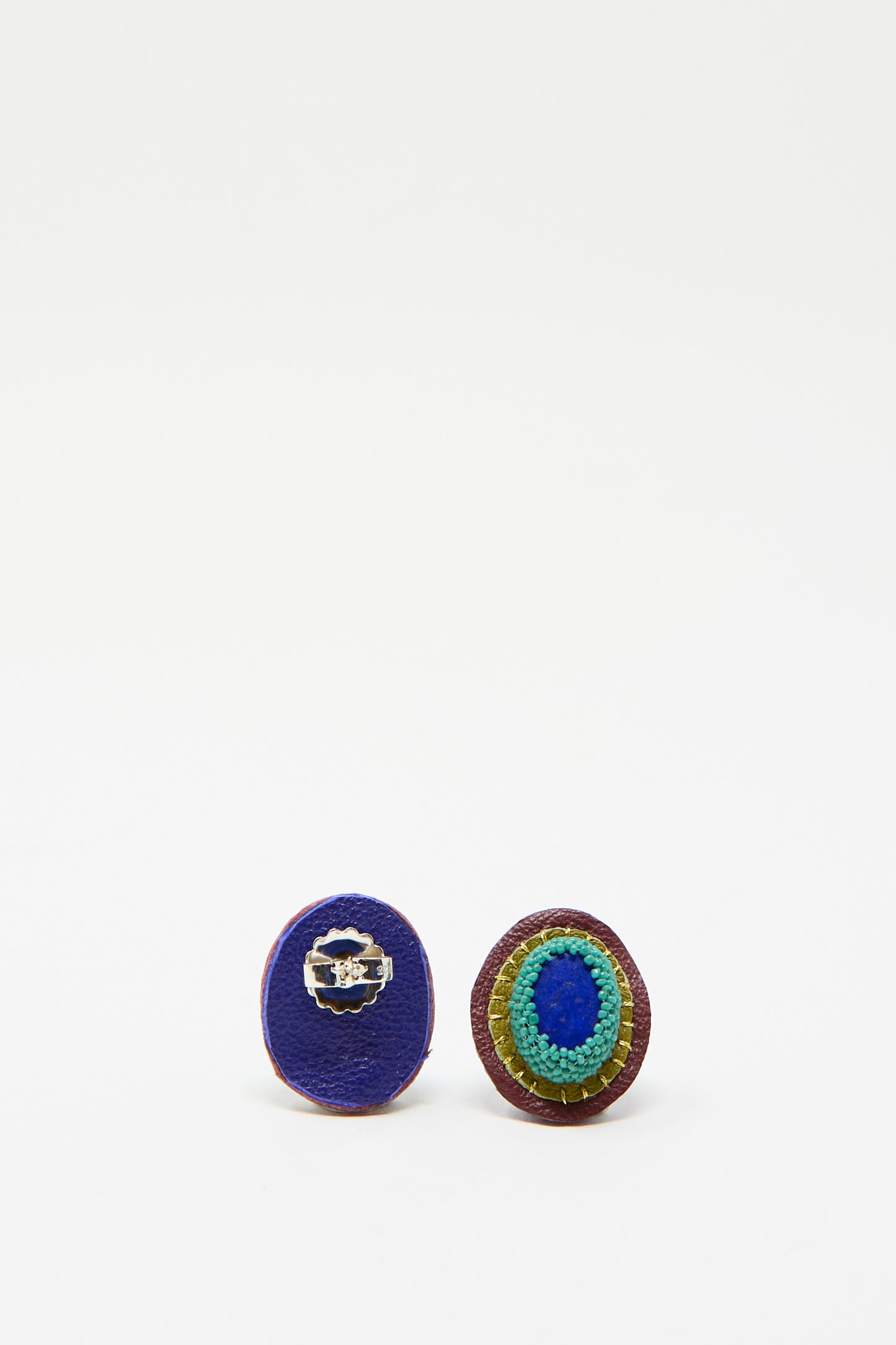 Two vibrant round earrings; one from the back featuring blue leather with a silver clasp, while the other displays layered stitching in blue, green, and yellow hues. Robin Mollicone's Oval Earrings in Lapis are seen against a white background.