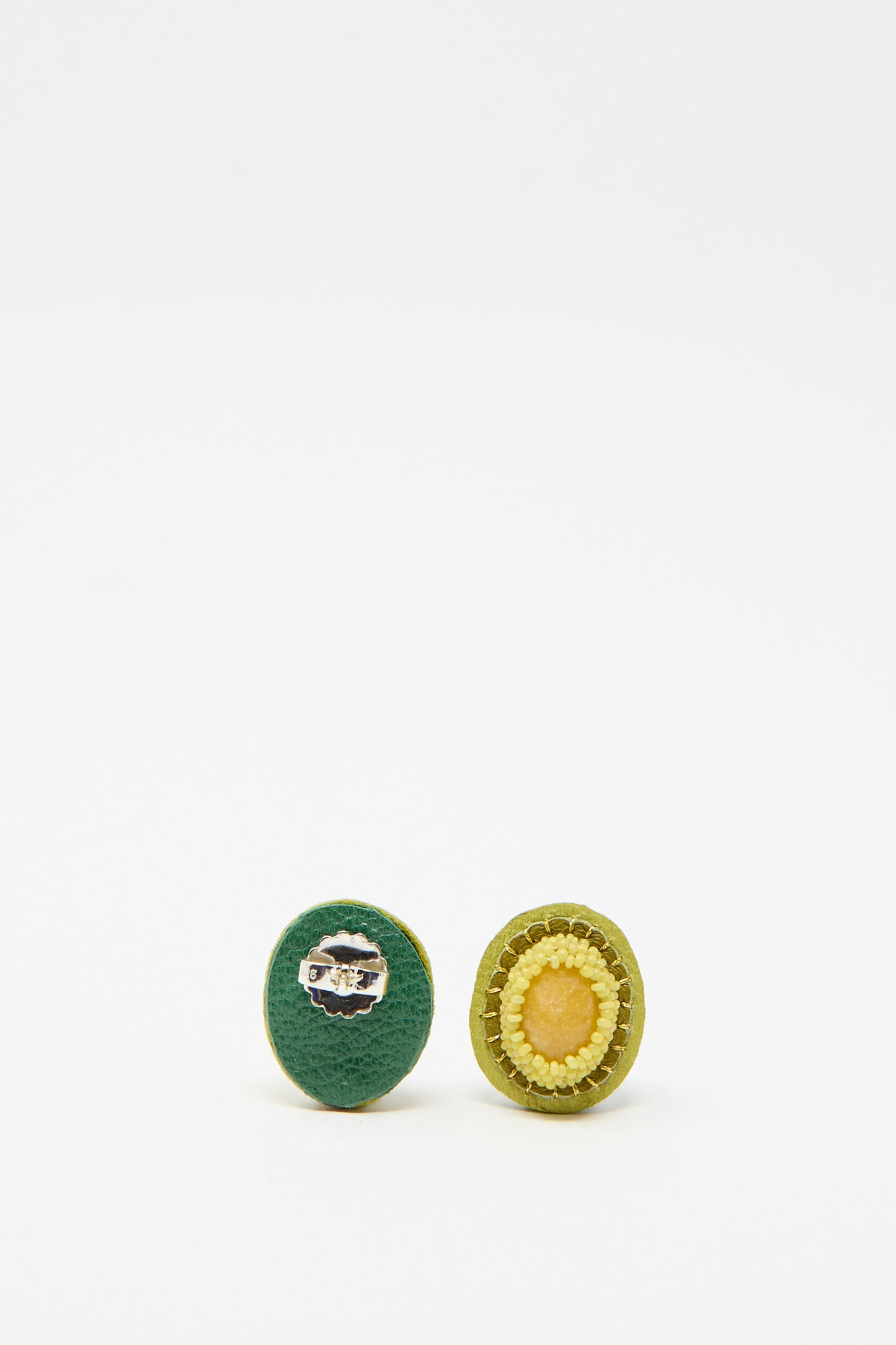 Oval studs by Robin Mollicone are [laced against a white background, one facing away with green fabric surface adorned with a silver clasp, while the other displays a stunning yellow stone.