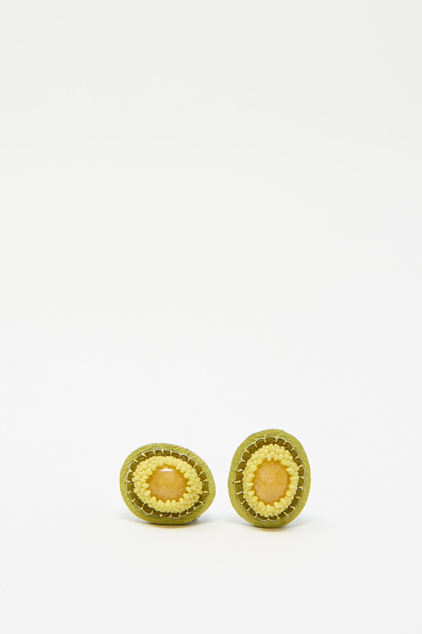 Robin Mollicone's Oval Earrings in Yellow Jasper resting against a white background, featuring green and yellow beading against a leather backing.