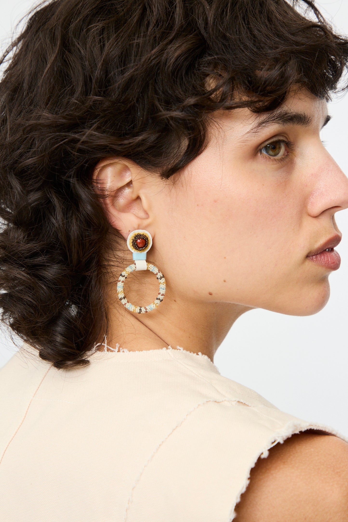 A person with short, dark curly hair wears a beige sleeveless top and Robin Mollicone's Small Beaded Hoop Earrings in Red Jasper, handmade in NYC.
