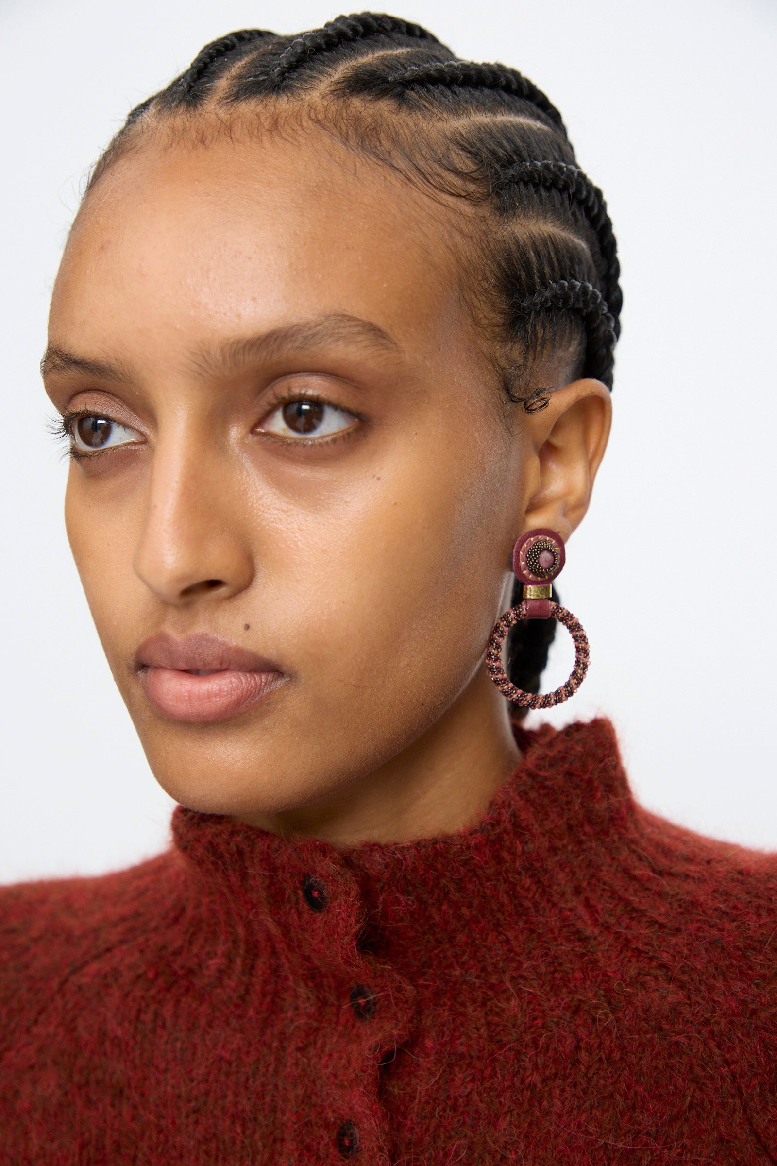 A woman with braided hair wears a textured burgundy top and Robin Mollicone’s Small Beaded Hoops in Pink with Rhodonite, gazing slightly to the side against a plain background.