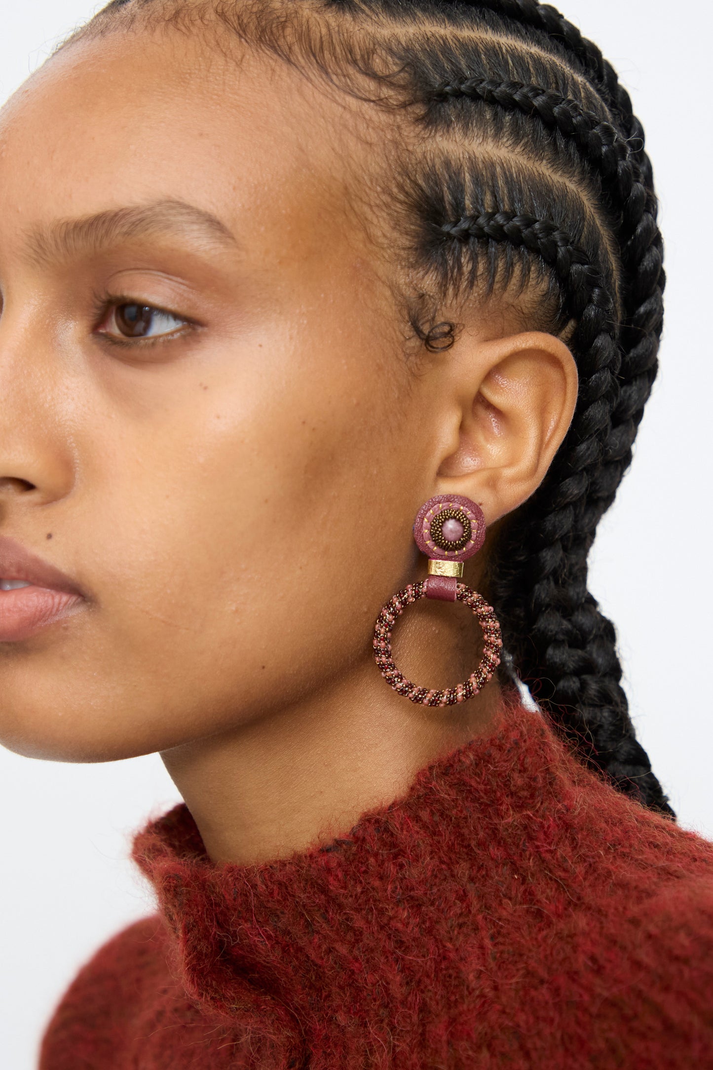 A person with braided hair wears a textured red sweater and Handmade in NYC Robin Mollicone's Small Beaded Hoops in Pink with Rhodonite, featuring a large circular pink design with a smaller circle above it.
