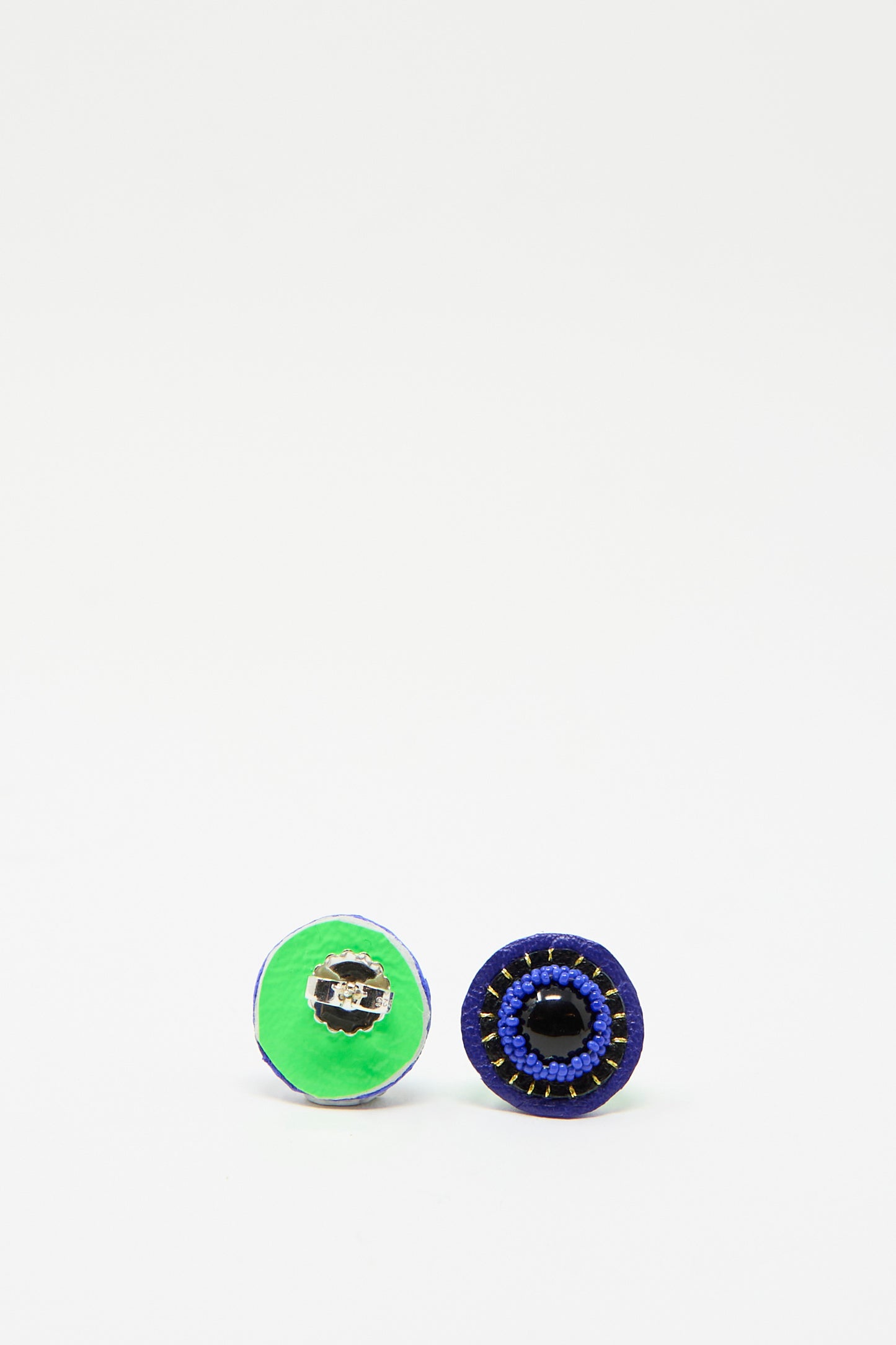 Displayed against a plain white background are two circular earrings by Robin Mollicone; one is facing backward showing its green leather back, and the other showcases its intricate handmade blue beaded design.