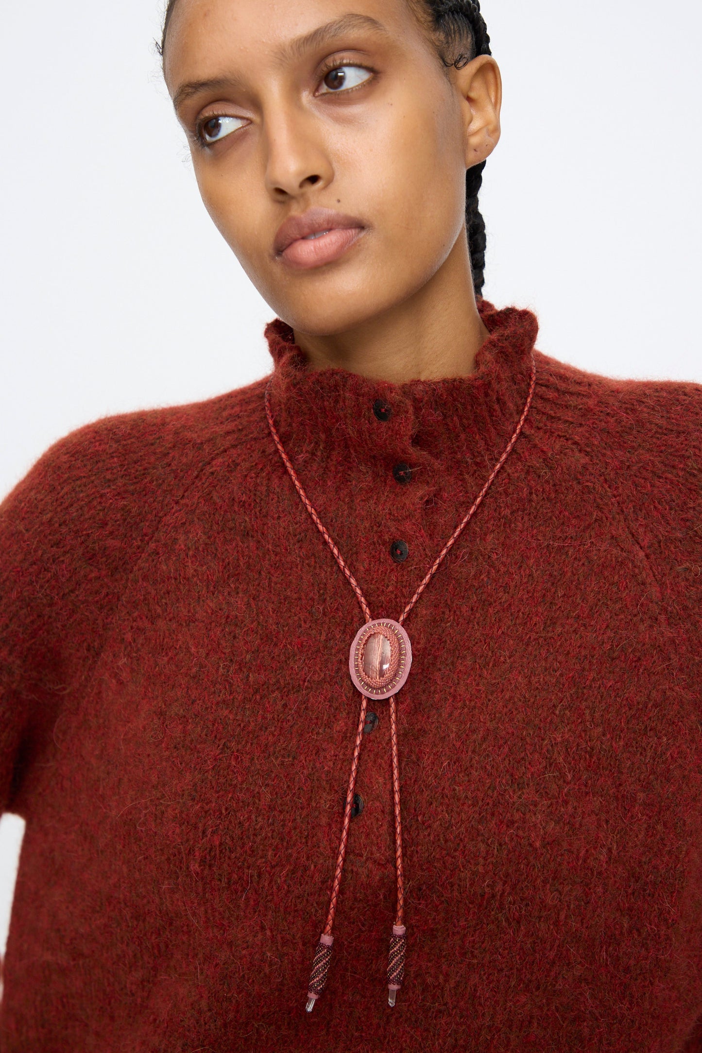 A person wearing a textured, dark red button-up sweater and braided hair, looking slightly to the side, styled with the Robin Mollicone Bolo Necklace in Snakeskin Jasper and Quartz.