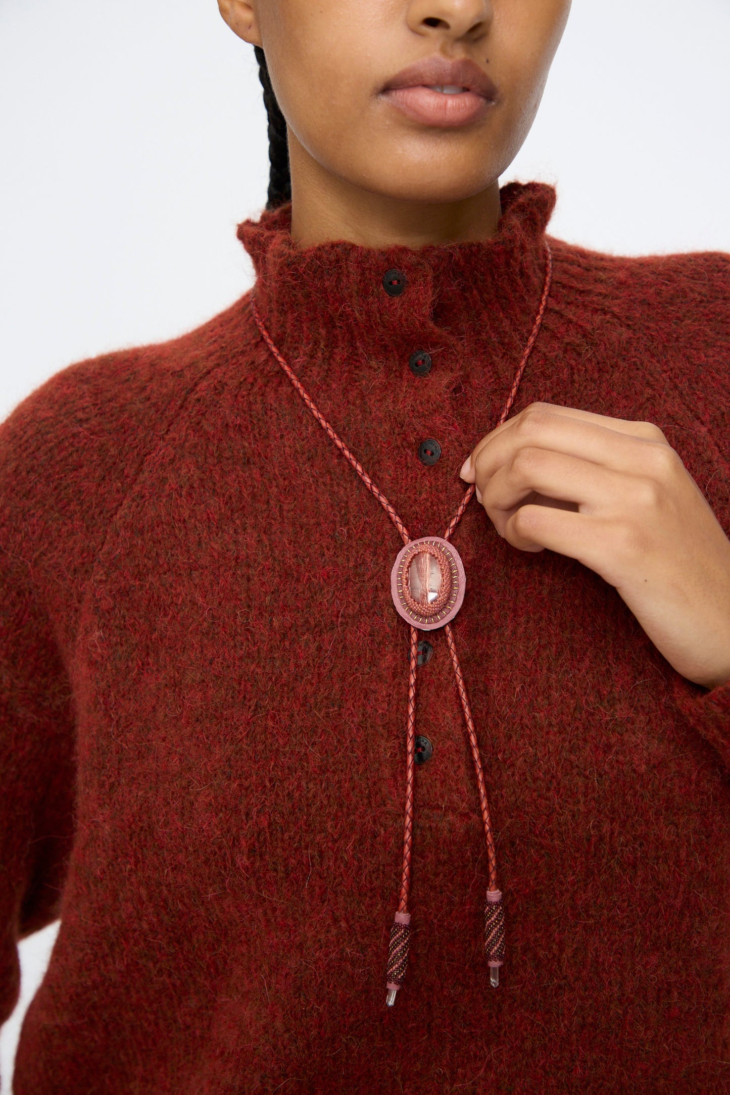 A person wearing a red sweater adjusts the Robin Mollicone Bolo Necklace in Snakeskin Jasper and Quartz, handmade in NYC, featuring a decorative oval stone and braided cords.