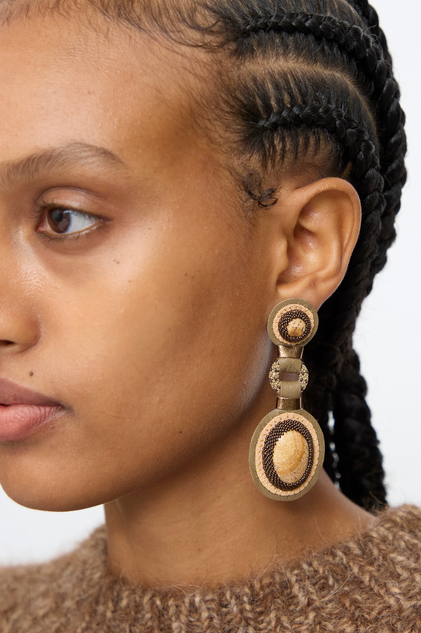 Wearing a brown sweater, a person with braided hair showcases Robin Mollicone’s Double Stone Earrings in Picture Jasper. Earth-toned, round earrings complement the look as only the left side of their face is visible.