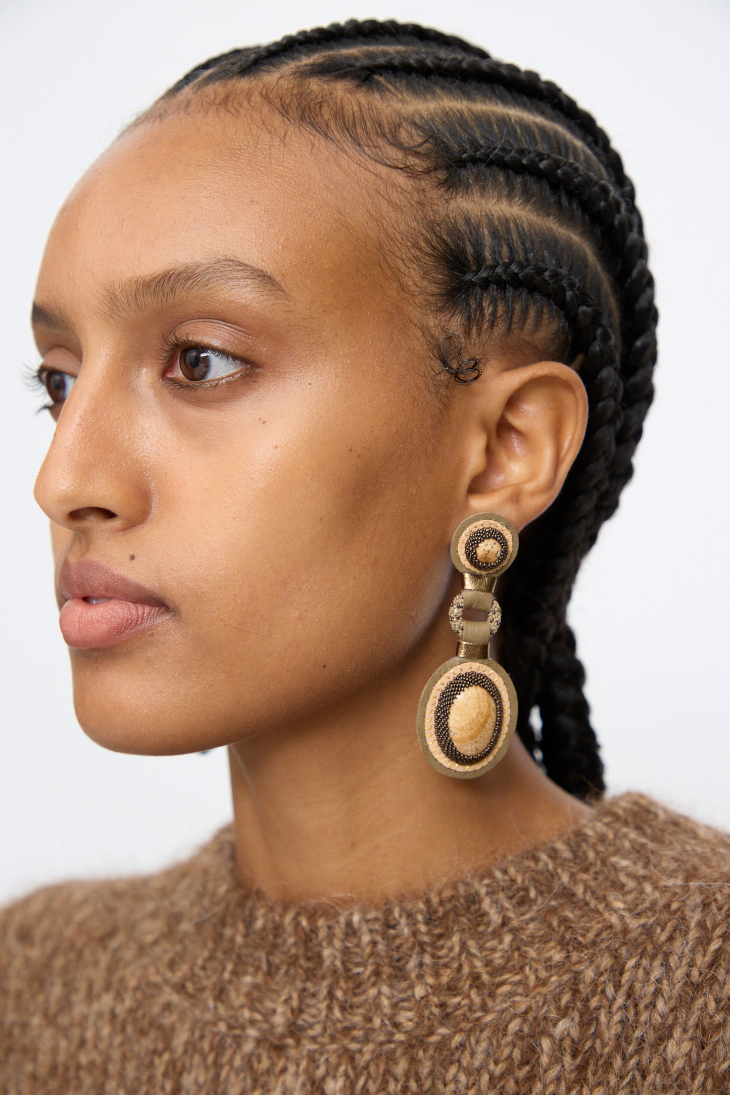 A person with neatly braided hair wears Robin Mollicone Double Stone Earrings in Picture Jasper and a brown knit sweater, facing left against a plain background.