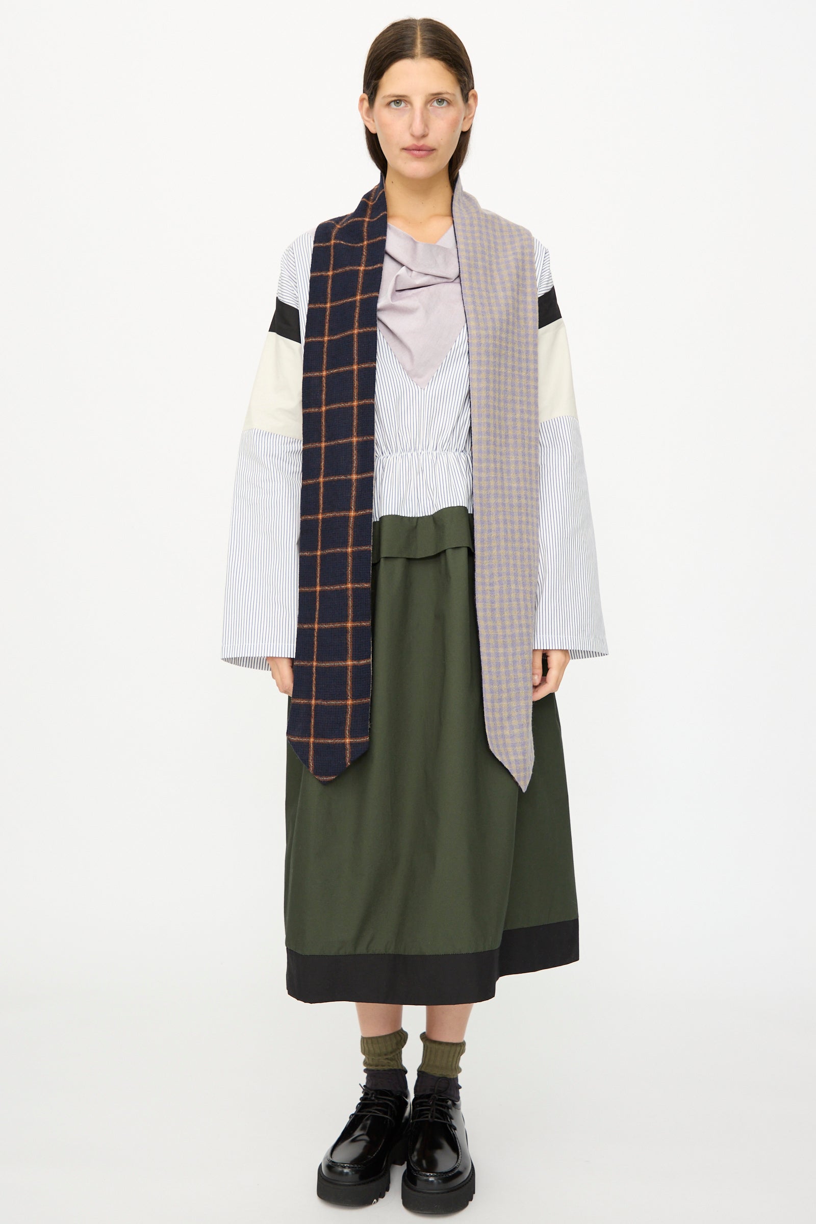 A woman faces forward against a plain white background, wearing a layered shirt, long dark skirt, black platform shoes, and the SC103 Check Wool Latch Scarf in Glossary.