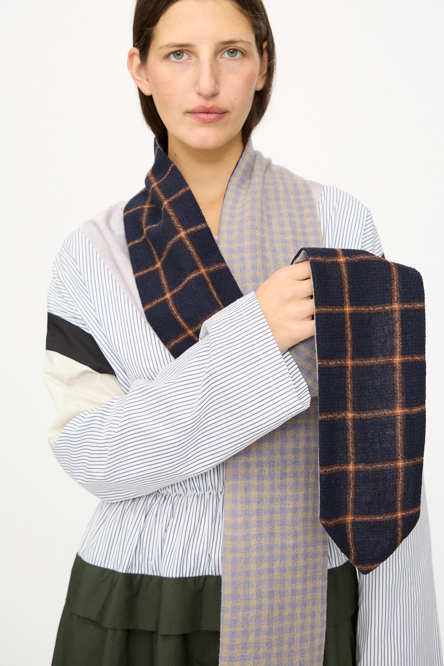 A person holds the SC103 Check Wool Latch Scarf in Glossary, featuring a navy base with blue and orange plaid, while wearing a loose, multi-patterned top against a light background.