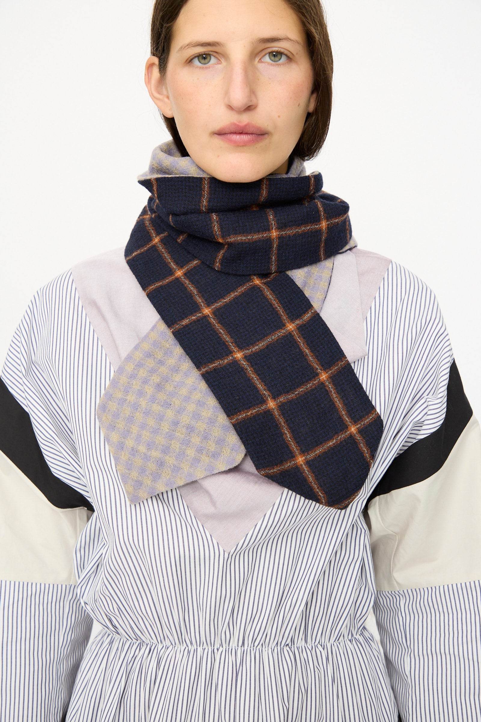 A person wears a striped dress paired with the SC103 Check Wool Latch Scarf in Glossary, featuring blue plaid and light checkered designs, wrapped around their neck.