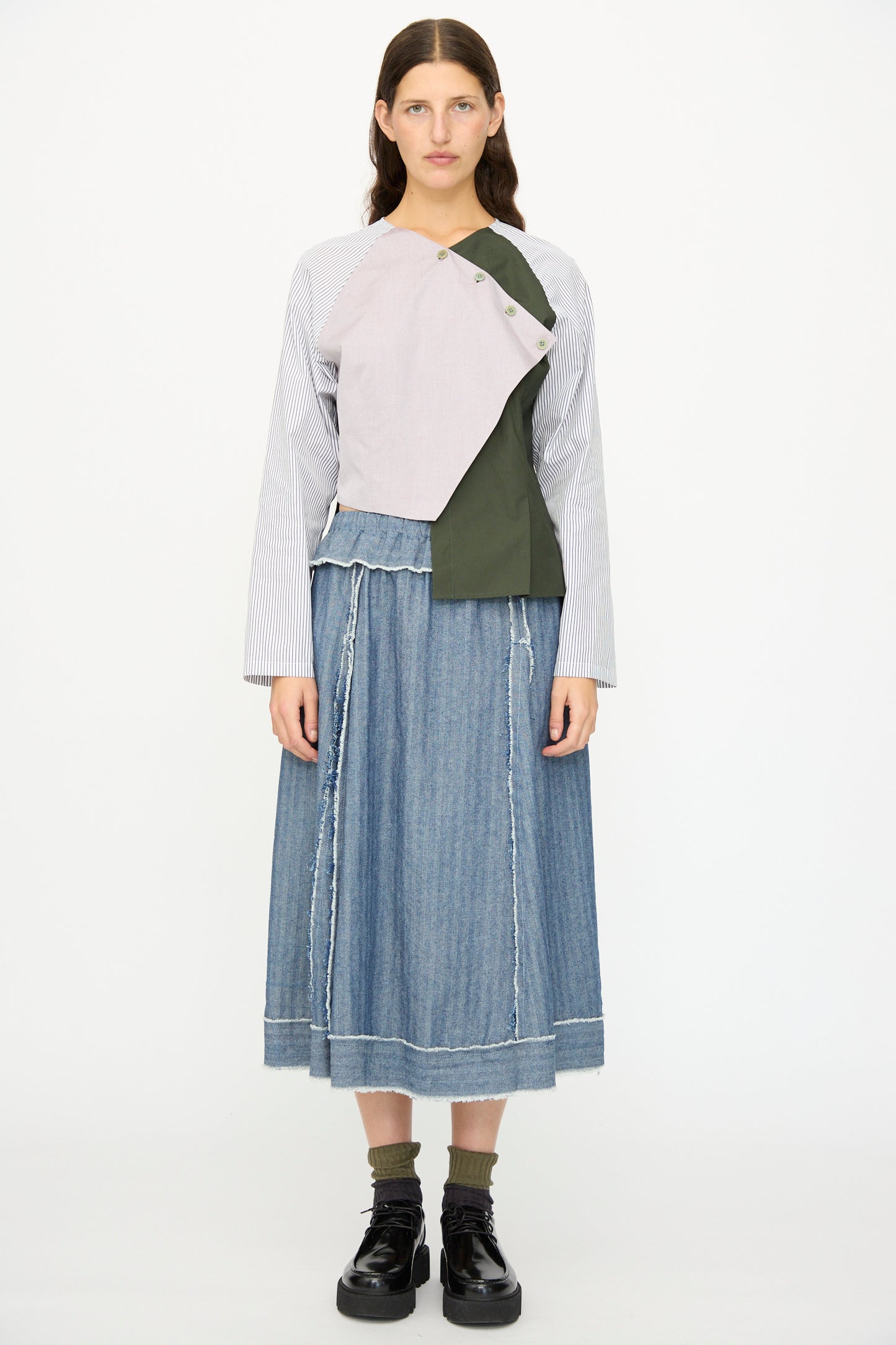 A woman faces forward wearing the SC103 Denim Radio Skirt in Herringbone, black boots, and a long-sleeve shirt with an asymmetrical design in gray, green, and striped fabrics.