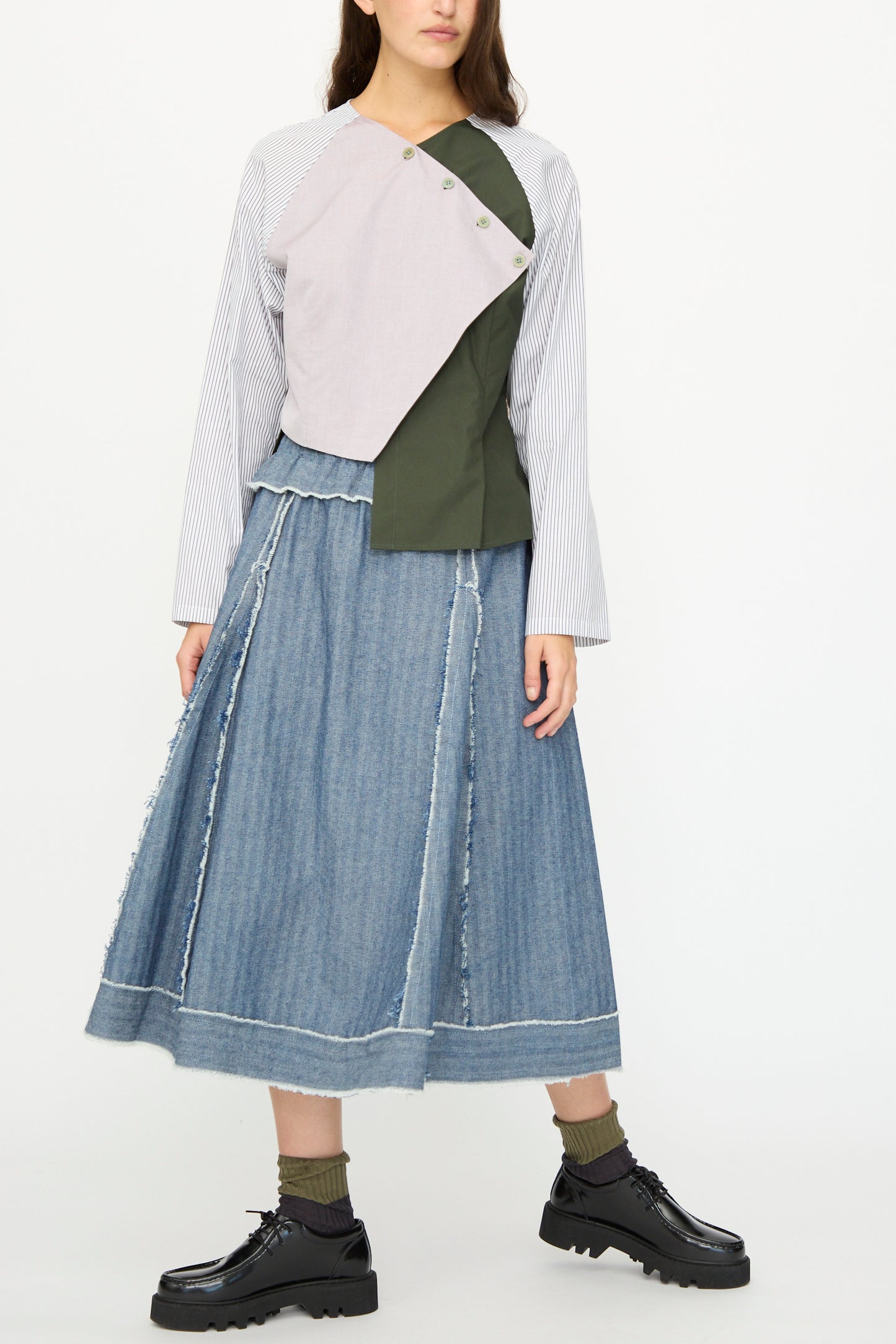 A woman wears SC103's Denim Radio Skirt in Herringbone, paired with black shoes, green socks, and a long-sleeve asymmetrical colorblock top in white, lilac, and olive green.