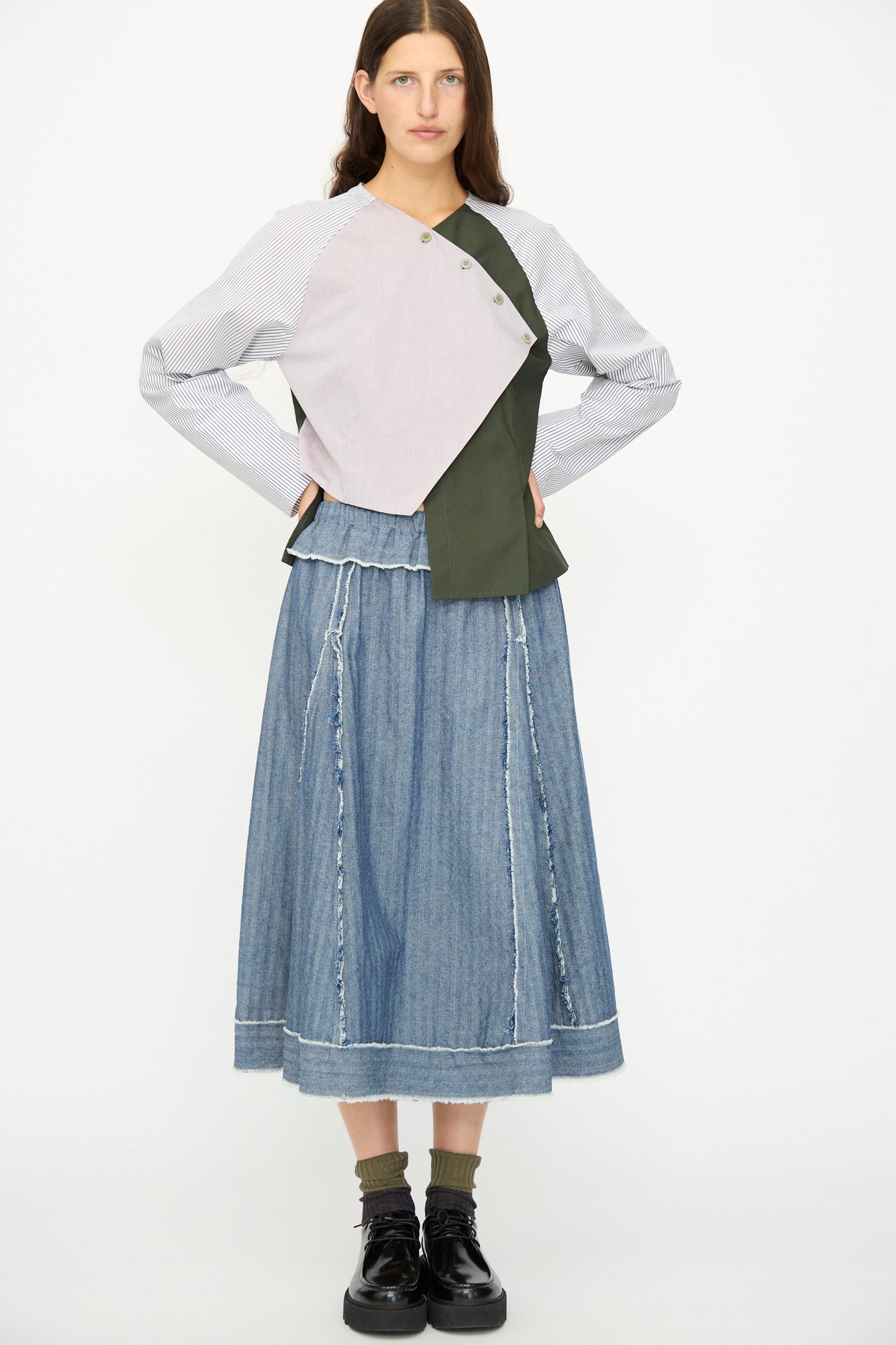 A woman stands against a white background wearing a patchwork long-sleeve top, green socks, black platform shoes, and the SC103 Denim Radio Skirt in Herringbone, a blue A-line midi skirt.