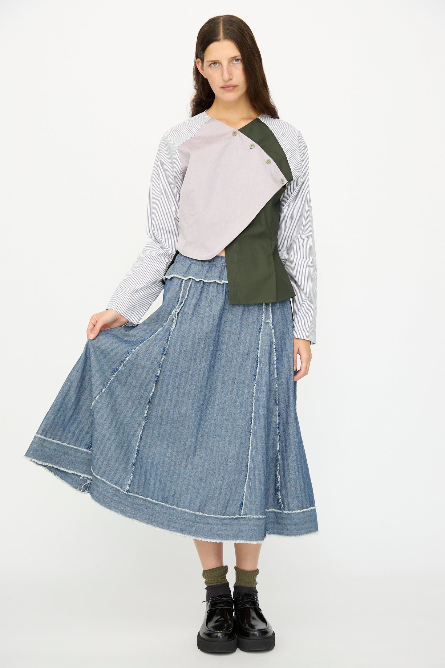 A woman stands against a white background wearing the SC103 Denim Radio Skirt in Herringbone, a patchwork top, socks, and black platform shoes.
