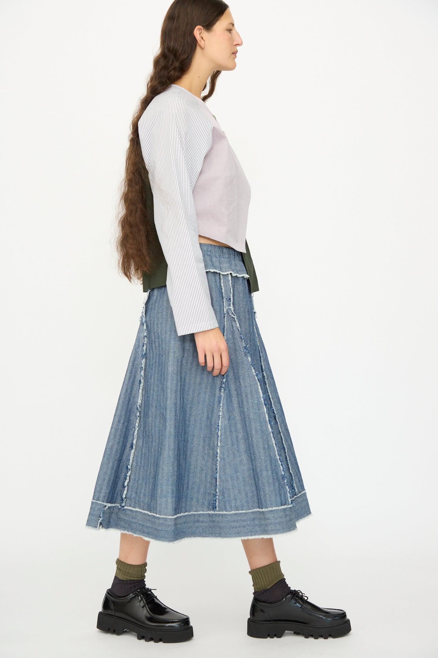 A woman with long brown hair wears a color-blocked long-sleeve top, green socks, black platform shoes, and the SC103 Denim Radio Skirt in Herringbone, standing against a plain white background.