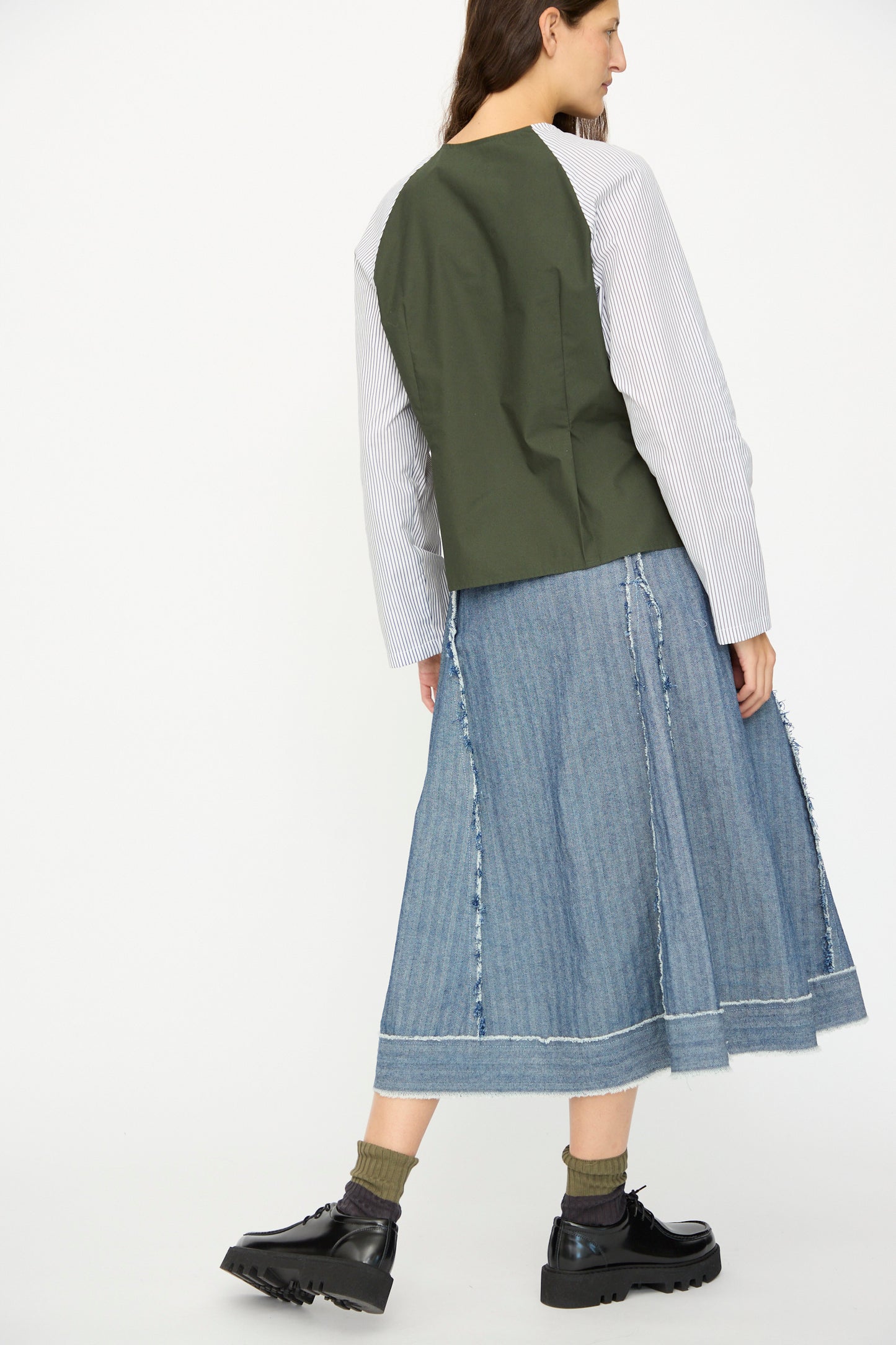 A person faces away, wearing a two-tone long sleeve shirt, green socks, black platform shoes, and the SC103 Denim Radio Skirt in Herringbone, a blue A-line midi skirt with raw edges.