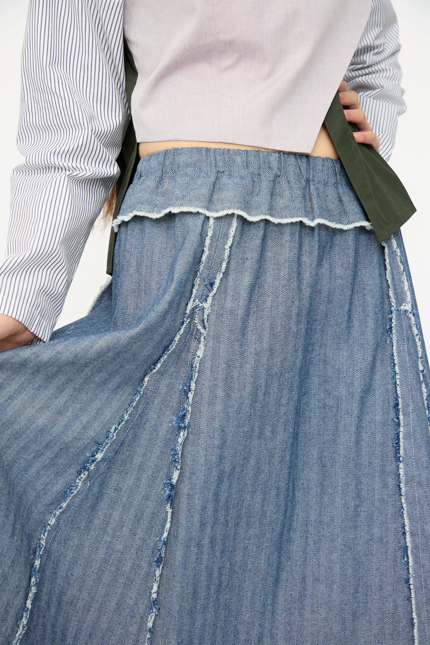 A person wears a light pink top with blue and white stripes on one sleeve and the SC103 Denim Radio Skirt in Herringbone, featuring a high-waisted A-line silhouette, visible seams, and a raw hem.
