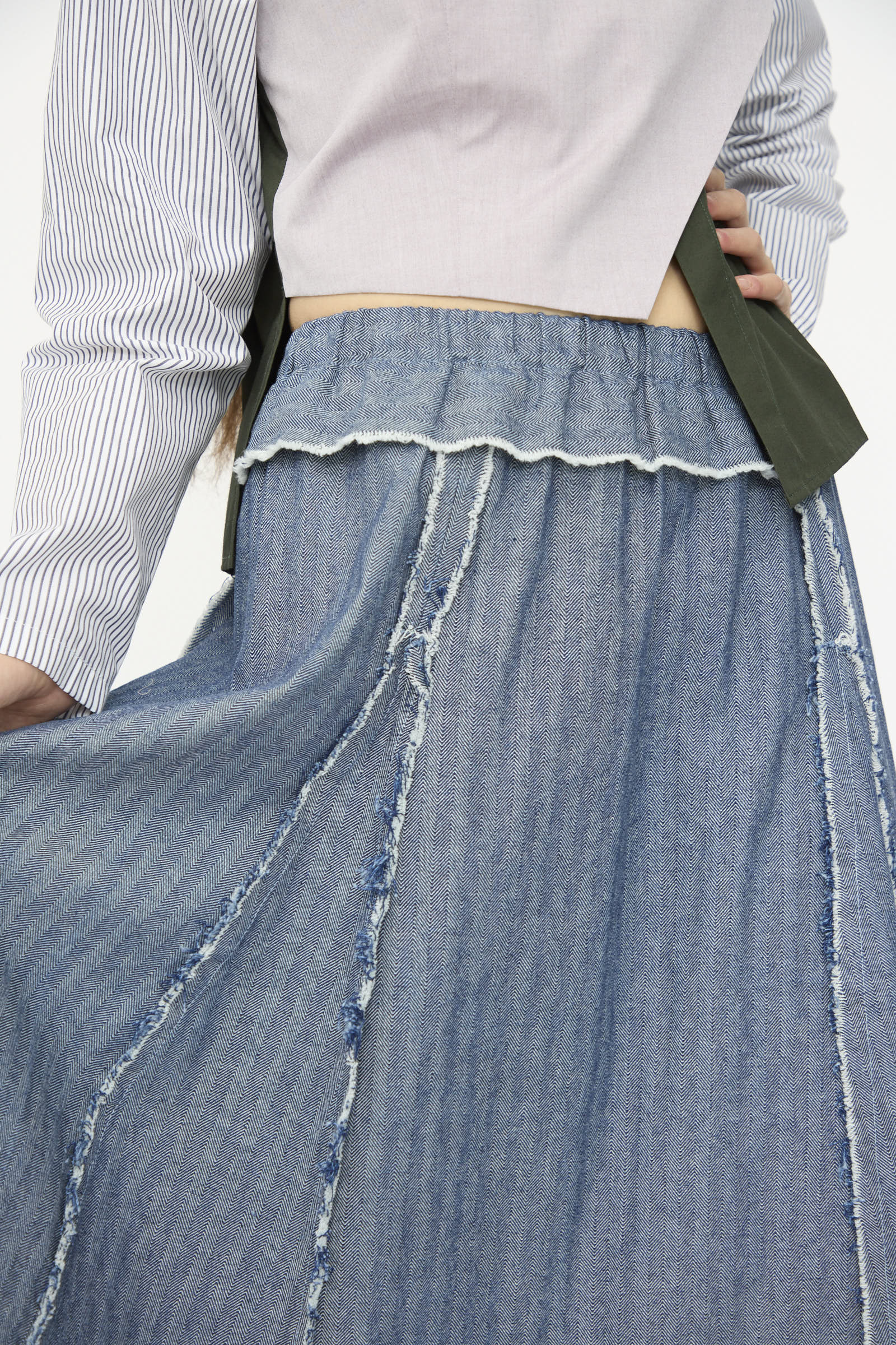 A person wears a light pink top with blue and white stripes on one sleeve and the SC103 Denim Radio Skirt in Herringbone, featuring a high-waisted A-line silhouette, visible seams, and a raw hem.