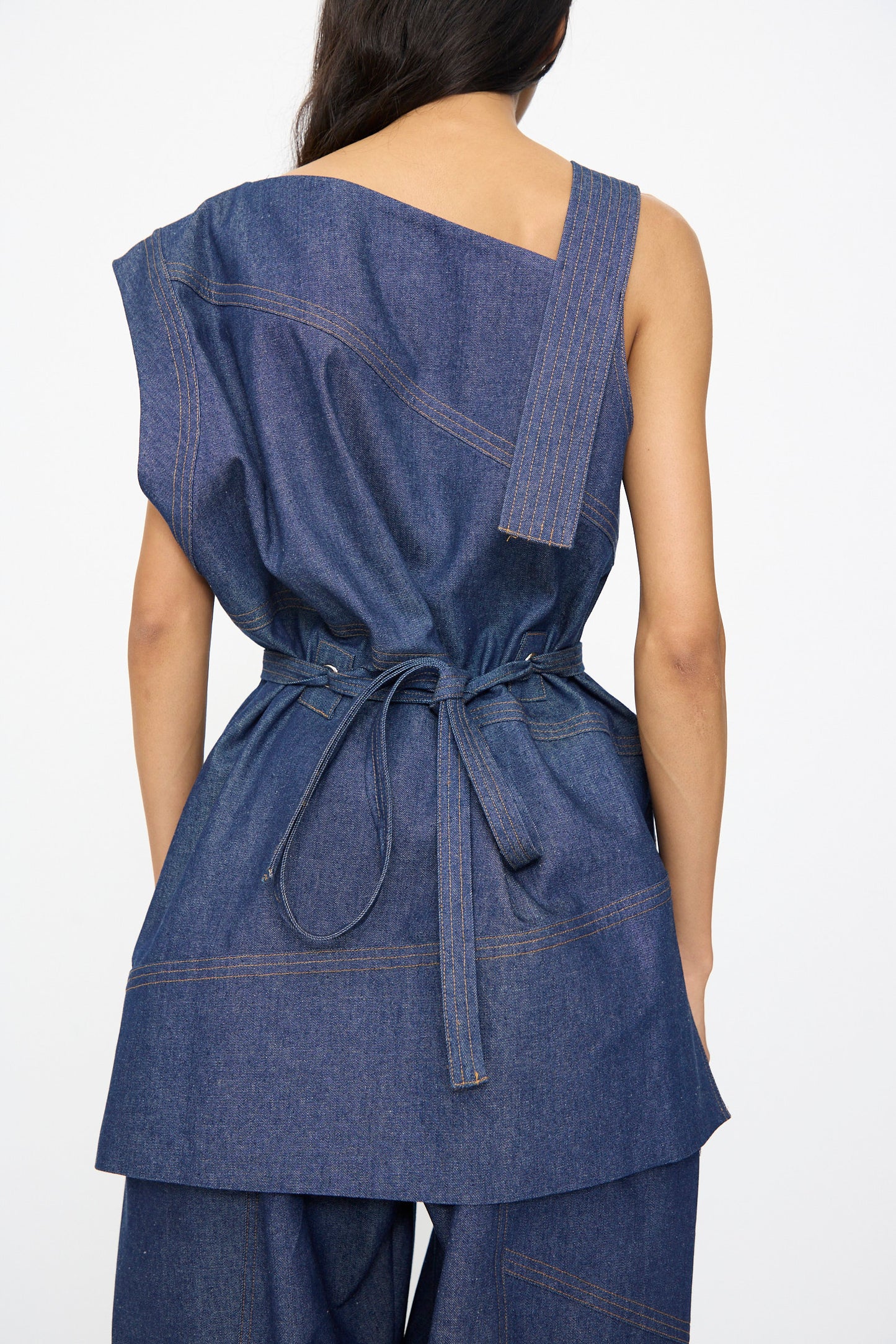 A woman is seen from the back wearing the SC103 Denim Sail Top in Soft Indigo, featuring a sleeveless design, asymmetrical neckline, and self-tie belt at the waist.