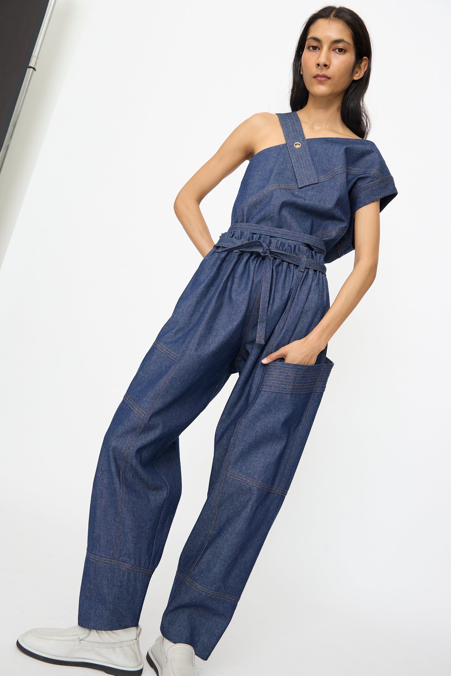 A woman stands against a white background wearing SC103's Denim Vault Pants in Soft Indigo—a relaxed fit, wide-leg style with a drawstring waist—paired with white shoes.