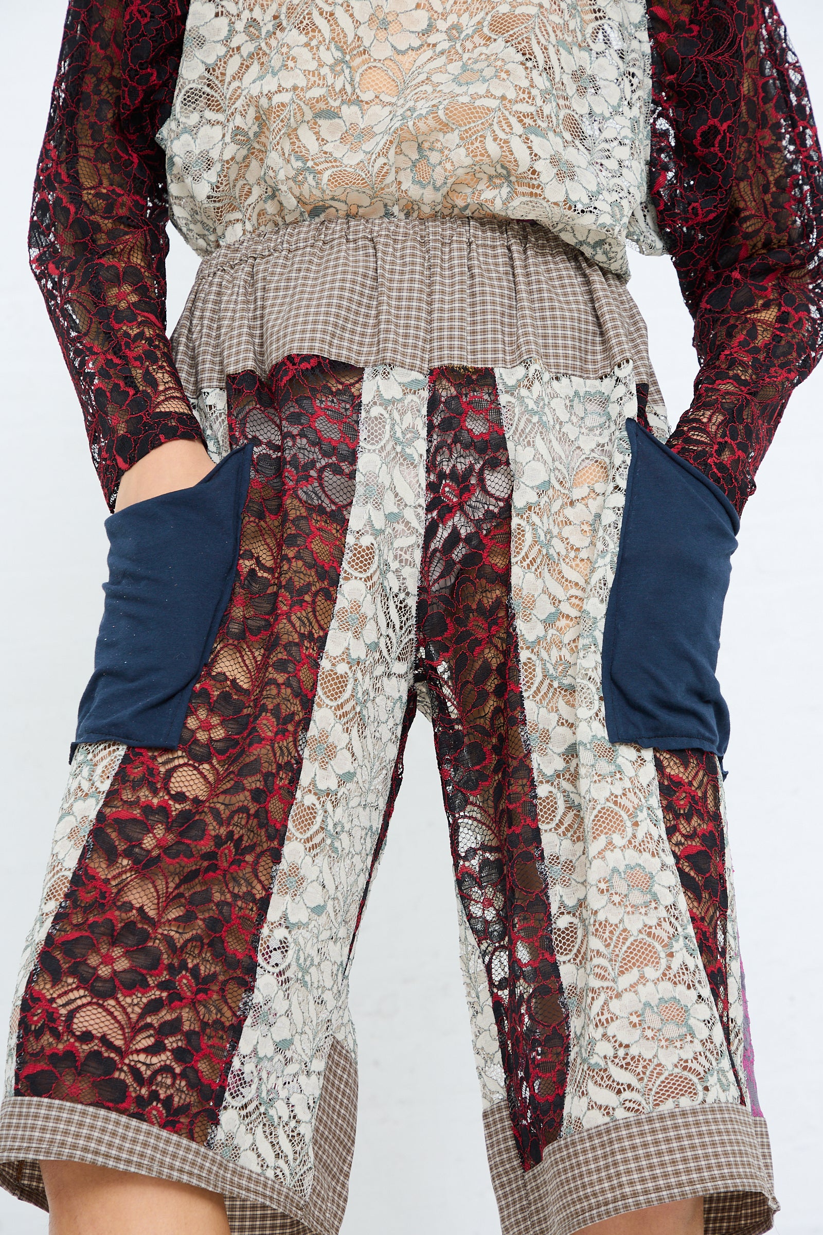 A person wearing an eclectic outfit with a patterned sleeve top, SC103 Lace Parallel Short in Thrash with an elasticated waist, and hands in pockets.