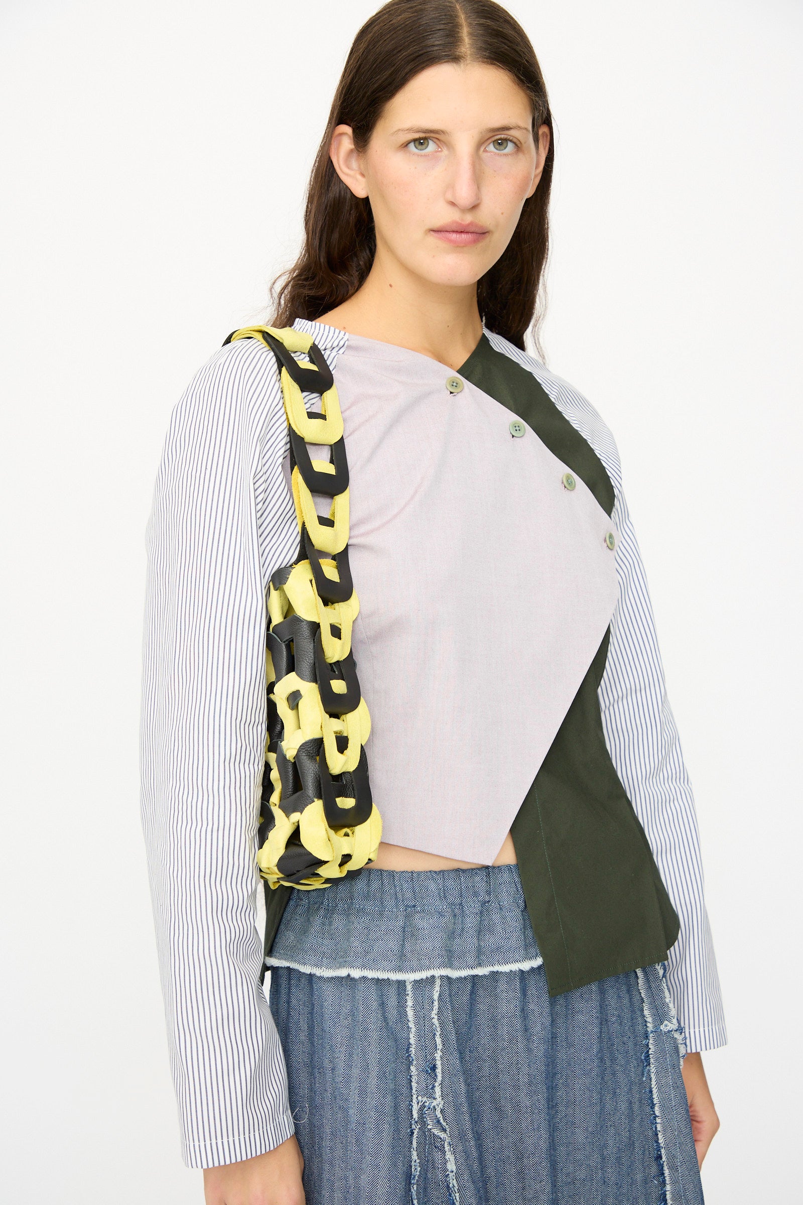 A woman with long brown hair wears blue jeans, an asymmetrical multi-colored top, and carries the SC103 Leather Links Tote in Dart—a handmade bag with interlocking links—against a plain background.