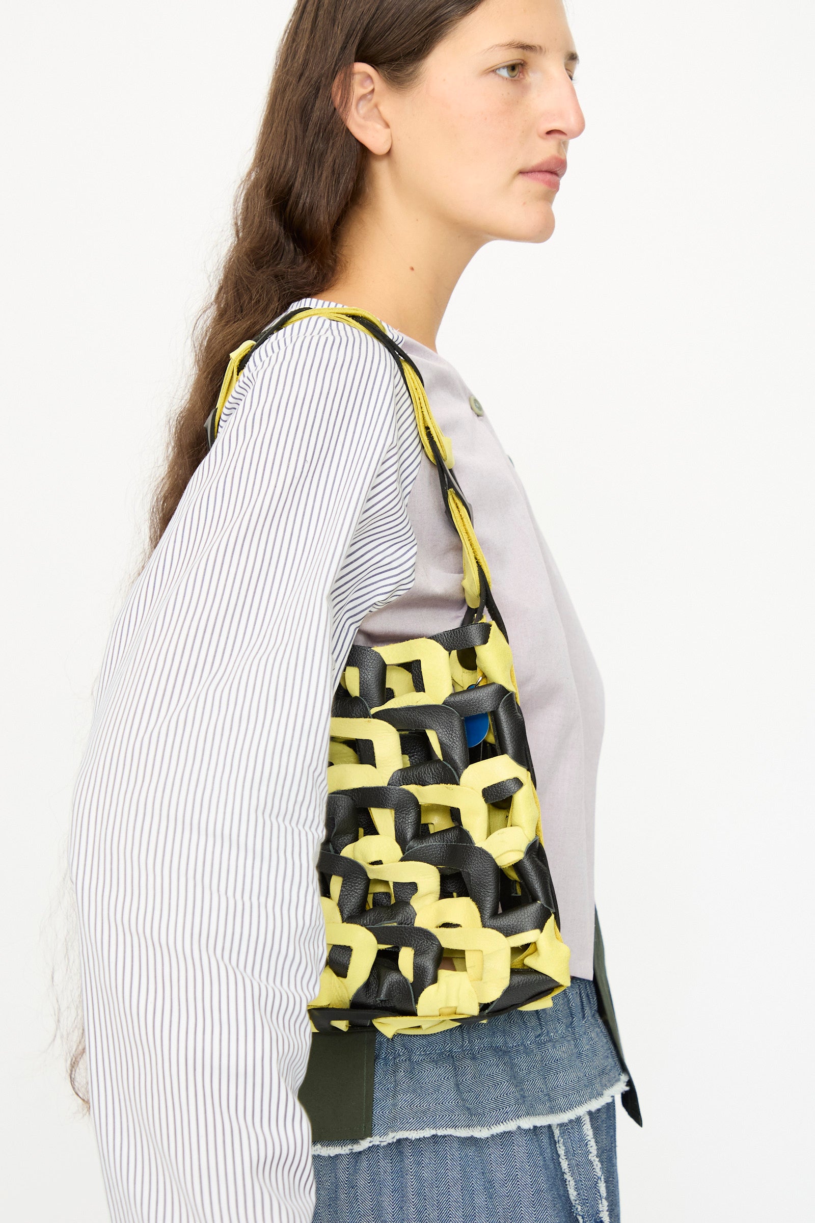 A woman in a long-sleeve shirt and jeans is shown in profile carrying the SC103 Leather Links Tote in Dart, a handmade bag with yellow and black interlocking links, over her shoulder.