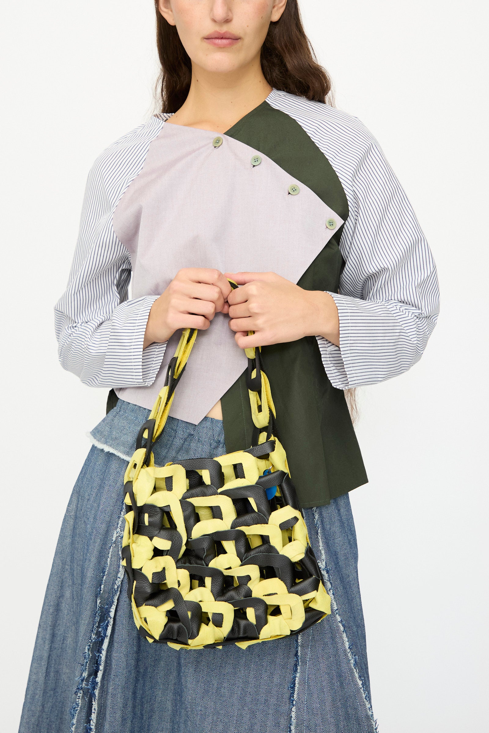 A person holds the SC103 Leather Links Tote in Dart, a handmade yellow and black bag with interlocking links, while wearing a patchwork top and distressed denim skirt against a plain white background.