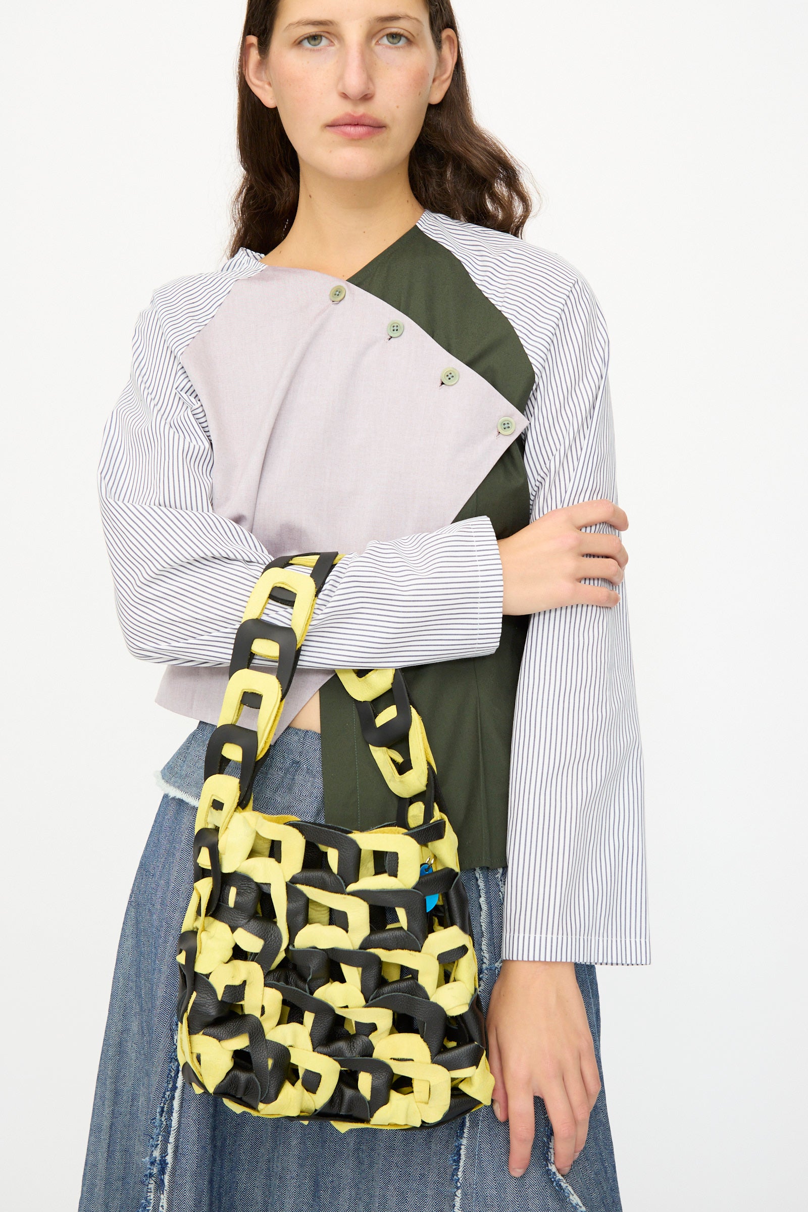 A woman holds the SC103 Leather Links Tote in Dart, featuring interlocking yellow and black links, while wearing a patchwork top and denim skirt against a plain white background.