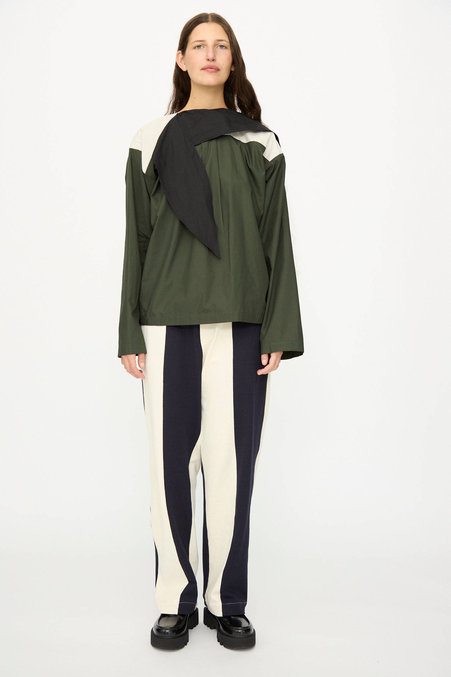 A person stands facing forward wearing the SC103 Poplin Compass Blouse in Relay, a color-blocked long sleeve green and black top, paired with two-tone black and white striped pants and black shoes.