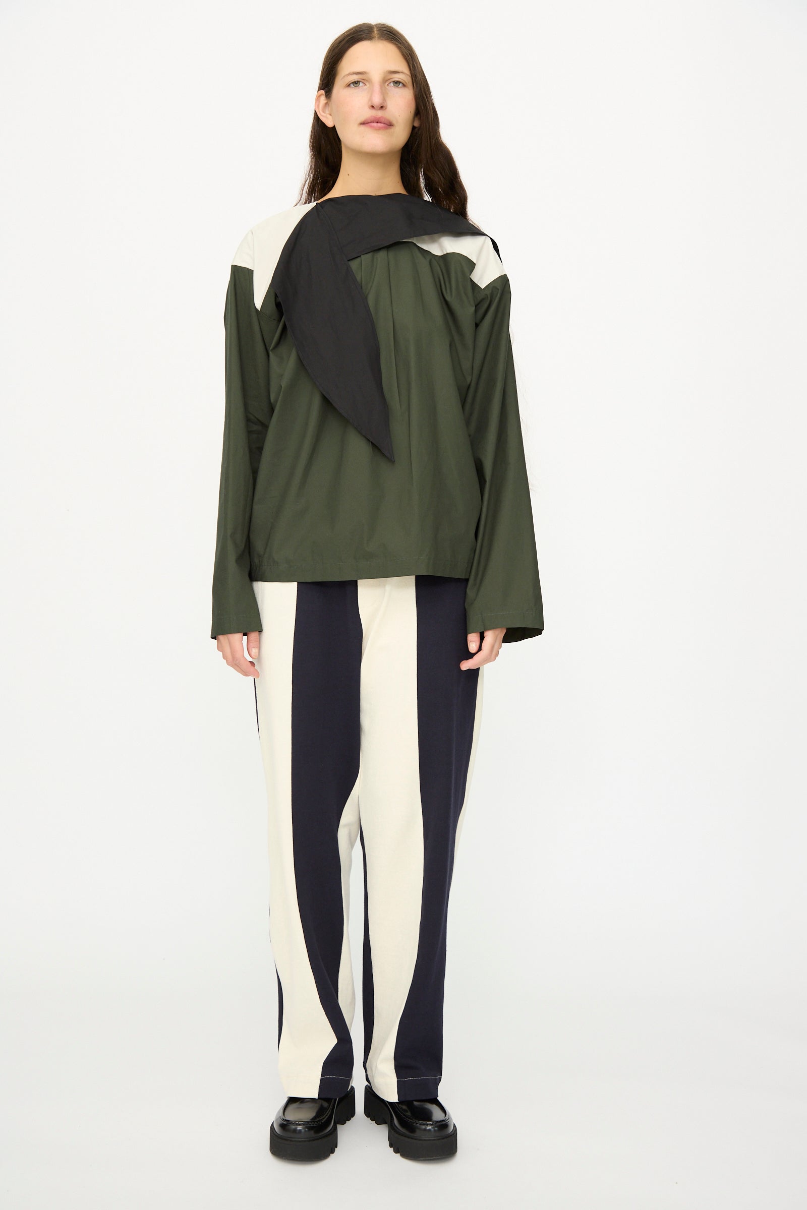A person stands facing forward wearing the SC103 Poplin Compass Blouse in Relay, a color-blocked long sleeve green and black top, paired with two-tone black and white striped pants and black shoes.