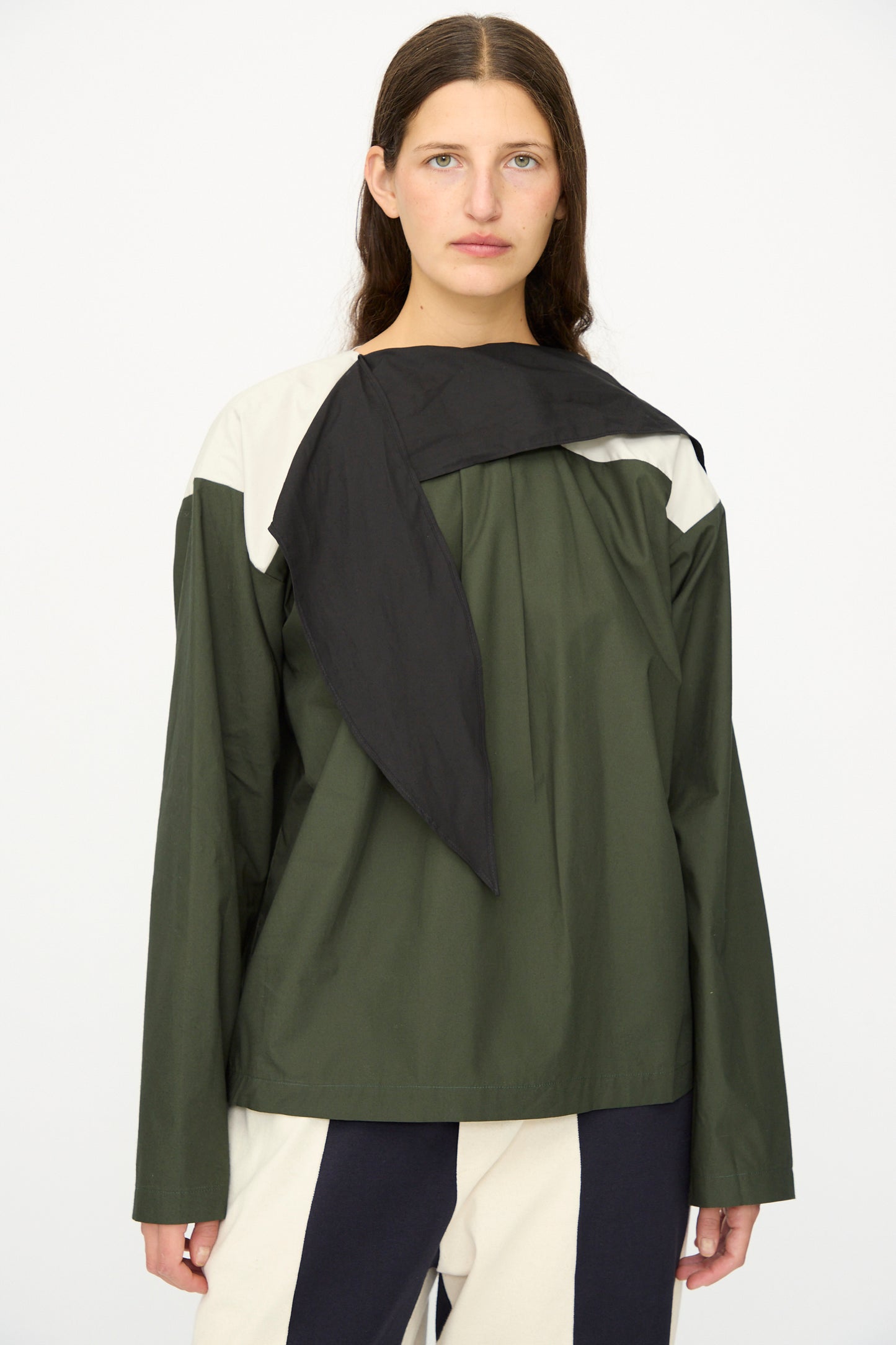 A person with long brown hair stands against a plain background, wearing the SC103 Poplin Compass Blouse in Relay, a green, black, and white color blocked top featuring a loose black scarf detail.