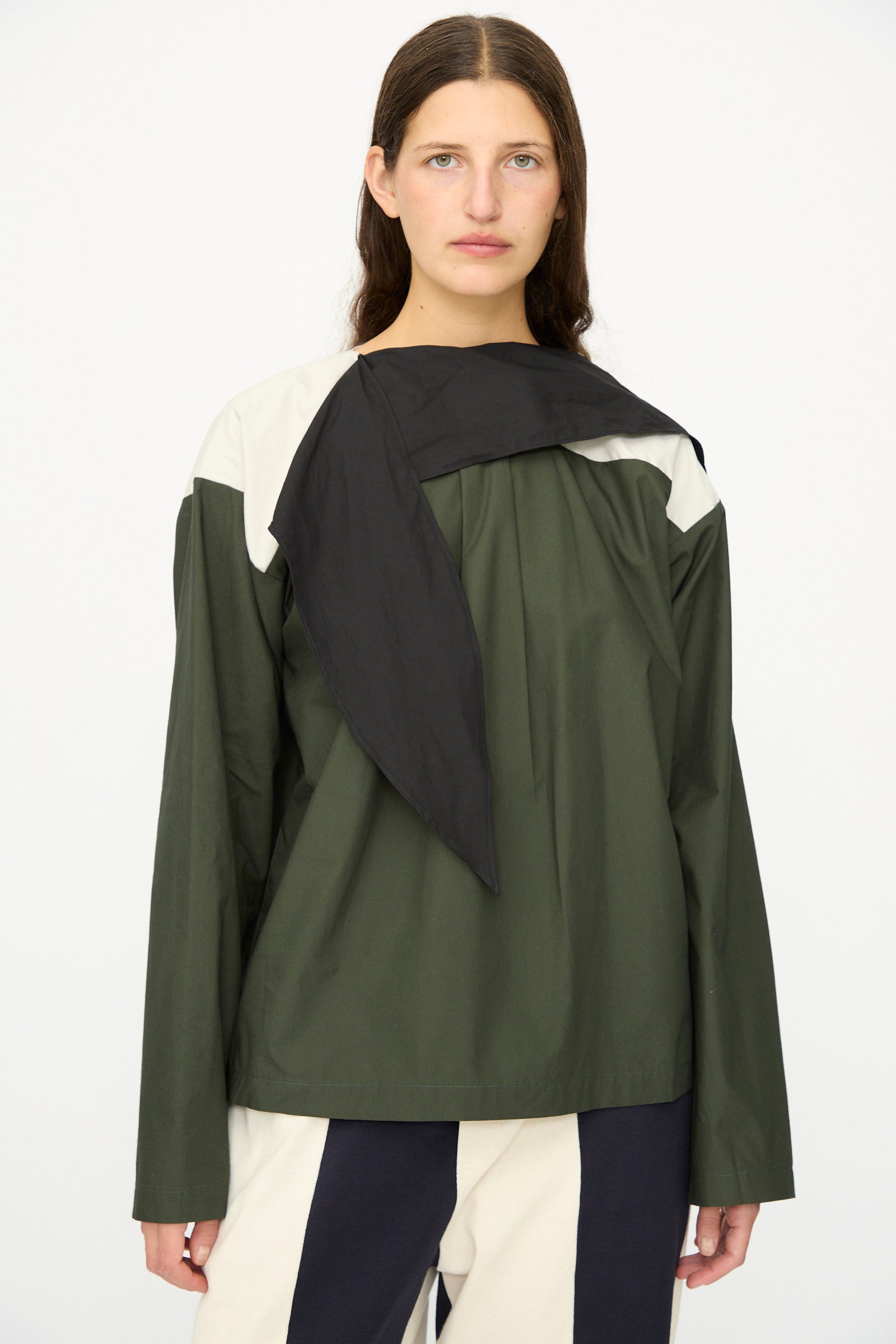 A person with long brown hair stands against a plain background, wearing the SC103 Poplin Compass Blouse in Relay, a green, black, and white color blocked top featuring a loose black scarf detail.