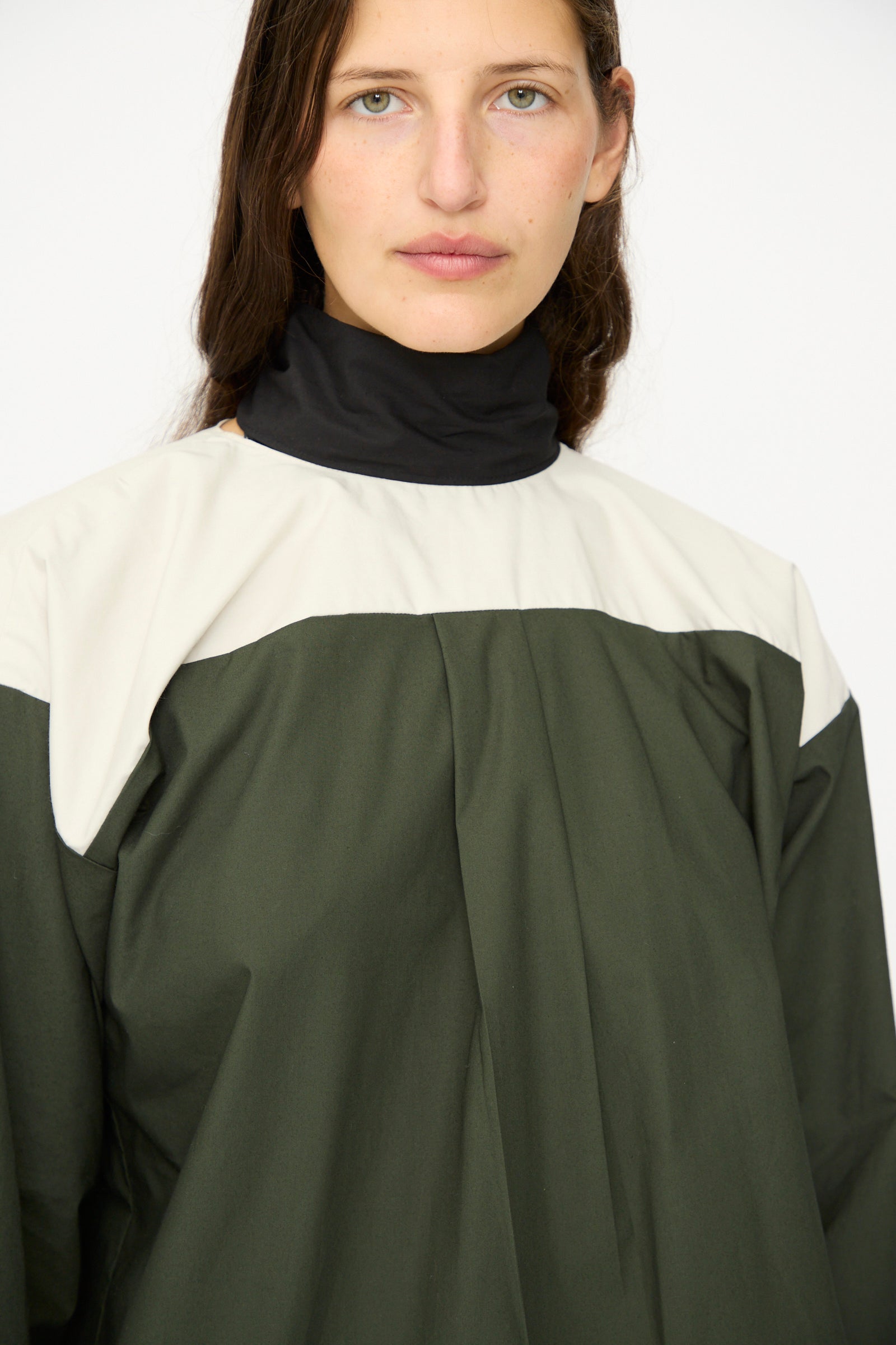 A person with long brown hair wears the SC103 Poplin Compass Blouse in Relay, a long-sleeve color-blocked top in dark green, white, and black cotton poplin, facing the camera against a plain background.