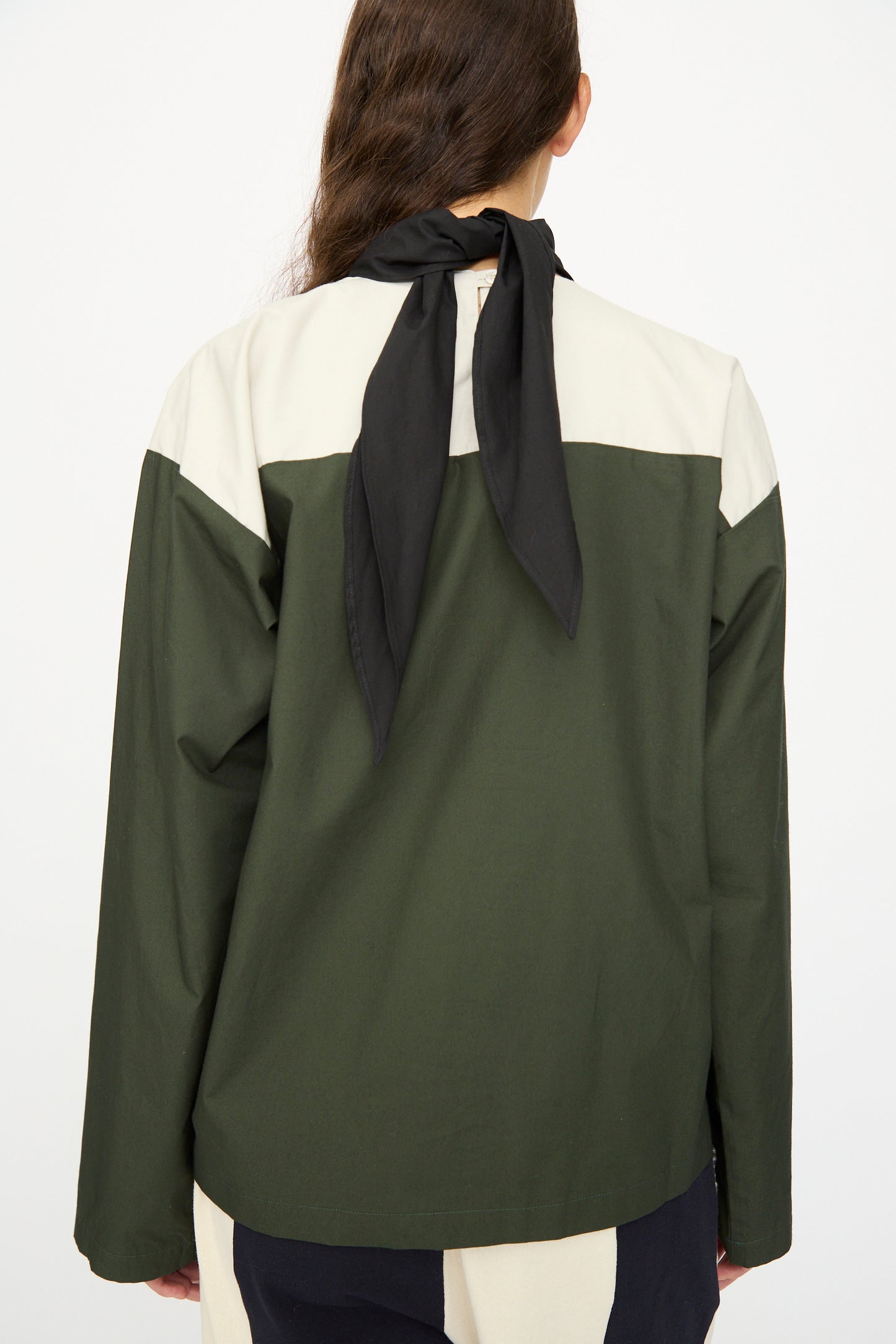 A person with long brown hair, seen from behind, wears the SC103 Poplin Compass Blouse in Relay, a color-blocked green and beige top, styled with a black scarf tied at the neck.